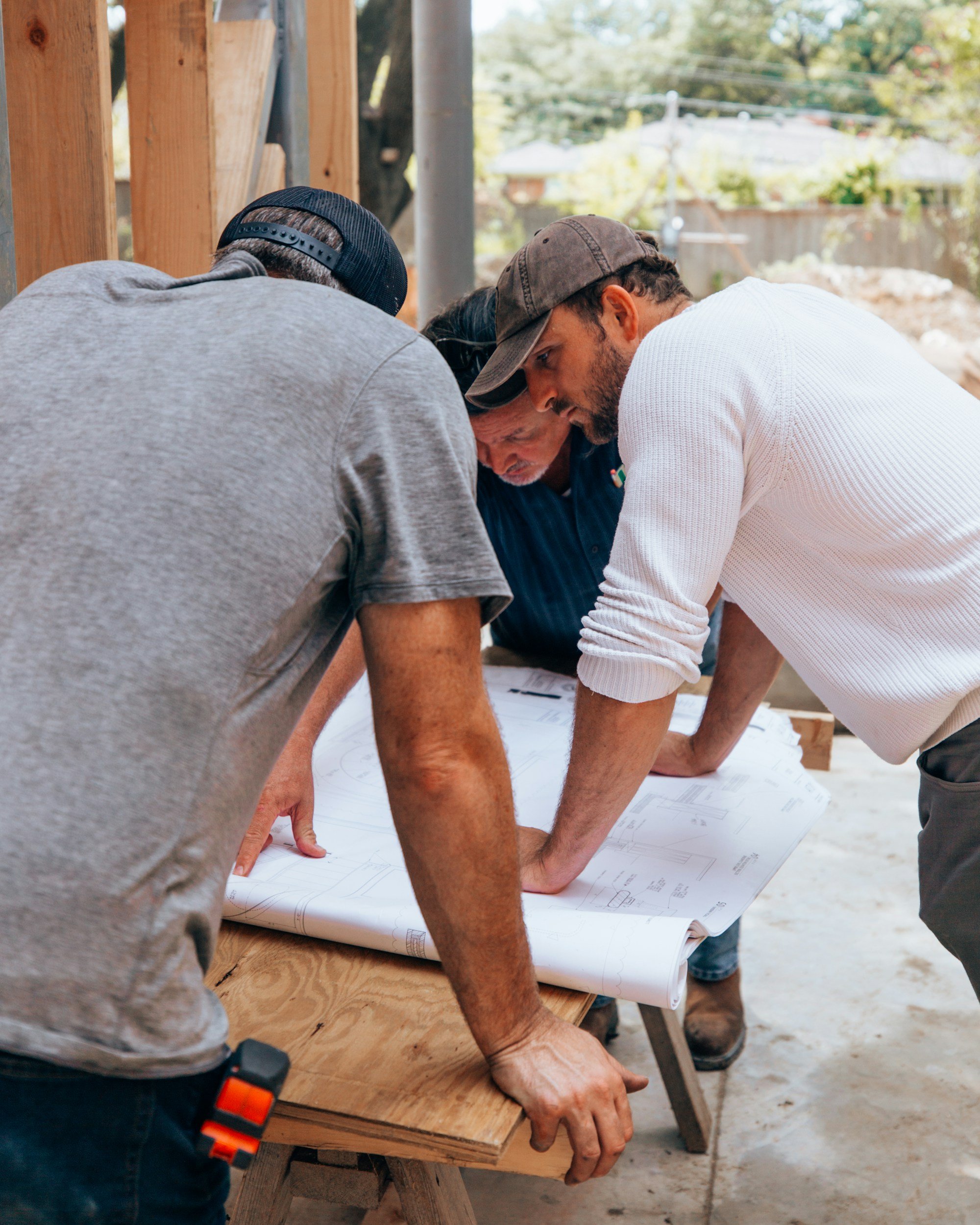 Three men are closely examining architectural blueprints spread out on a wooden work table at an outdoor construction site, with trees and houses visible in the background.