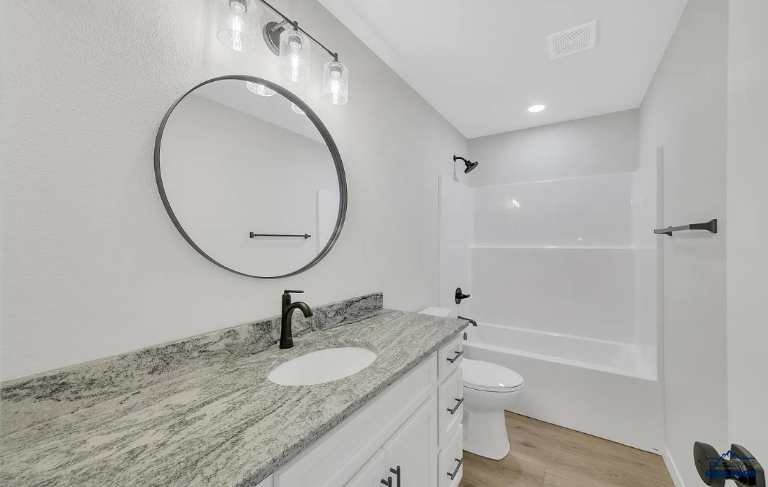 Modern bathroom with white vanity, granite countertop, oval mirror, black fixtures, bathtub with shower, white walls, and wood-look flooring.