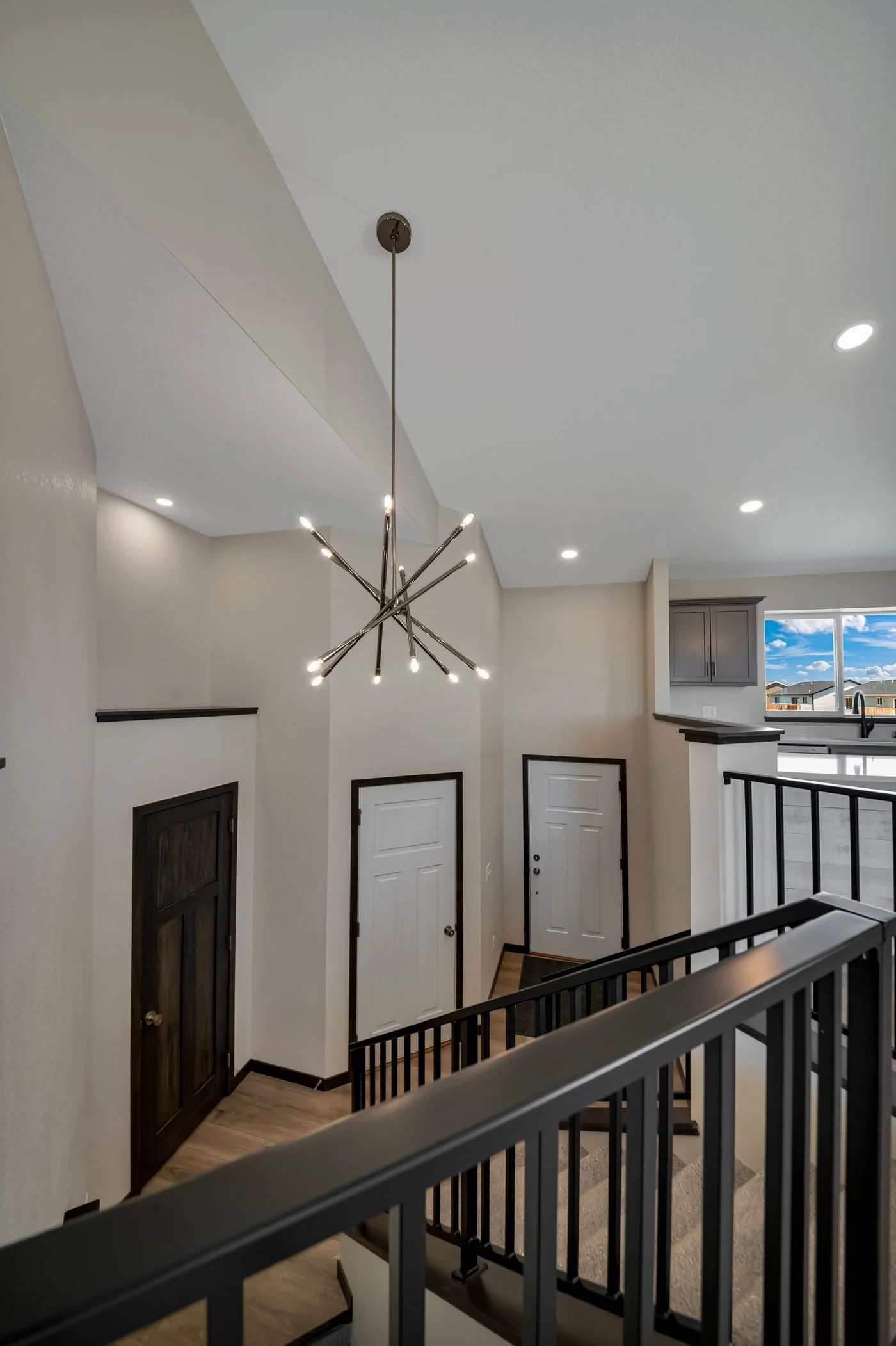 Interior of a modern house with a staircase, front door, and a kitchen window showing a blue sky and houses outside. A stylish chandelier hangs from the ceiling.