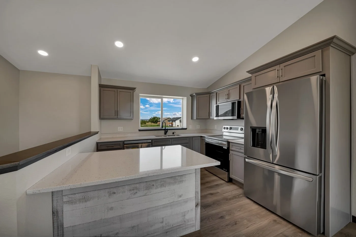 Modern kitchen with gray cabinets, stainless steel refrigerator, microwave, oven, and a large window overlooking a suburban neighborhood.