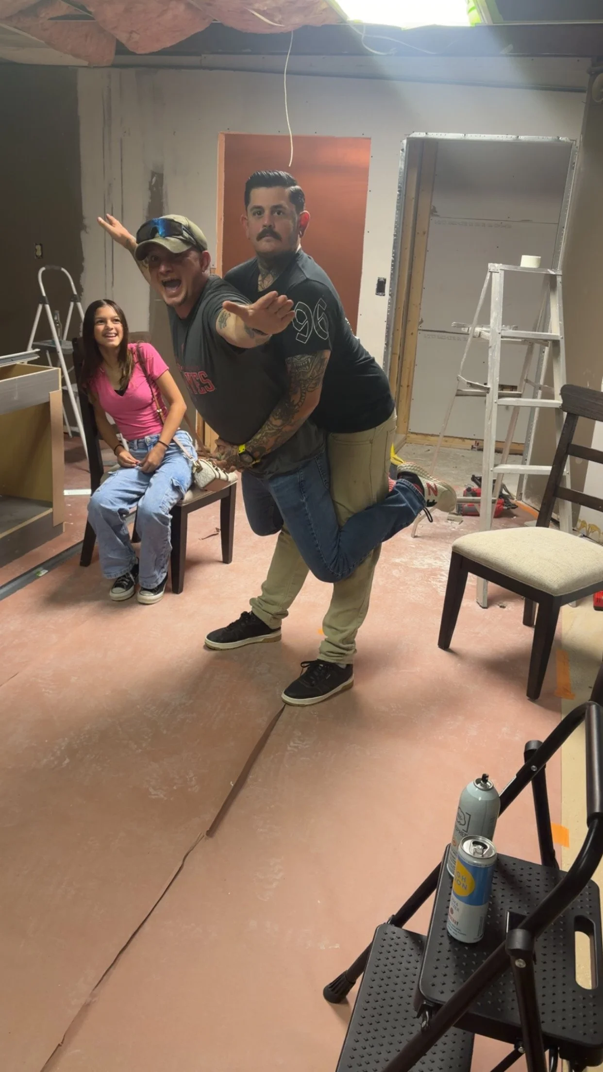 Two men are posing playfully in what appears to be a room under construction. One man is kneeling with his leg slightly raised, while the other leans over him, extending his arms. A young woman is seated on a chair to the left, smiling. The room has unfinished walls, ladders, and construction tools, with some seats and a water bottle visible.
