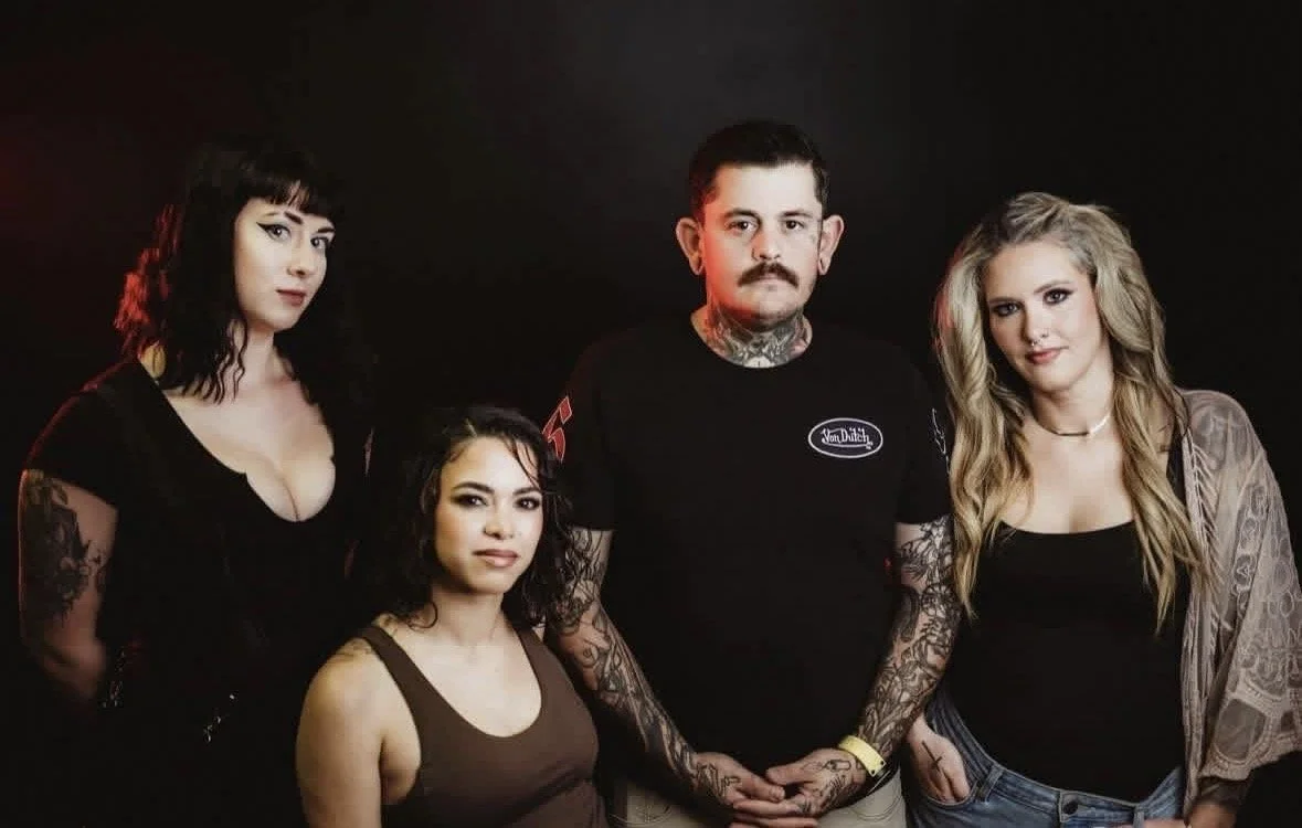 Group of four people standing against a dark background, two women with curly hair, one with black and one with blonde, and two men with mustaches, one with a tattooed neck, all in casual clothing.