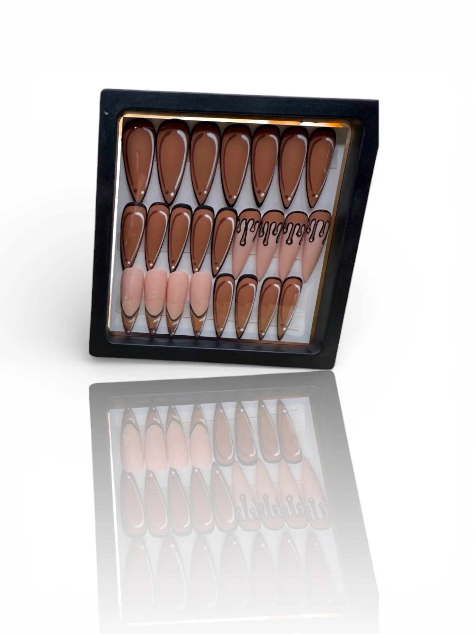 Brown almond press-on nails with black outline French tips, nude accent nails, and black drip nail art displayed in a black case.