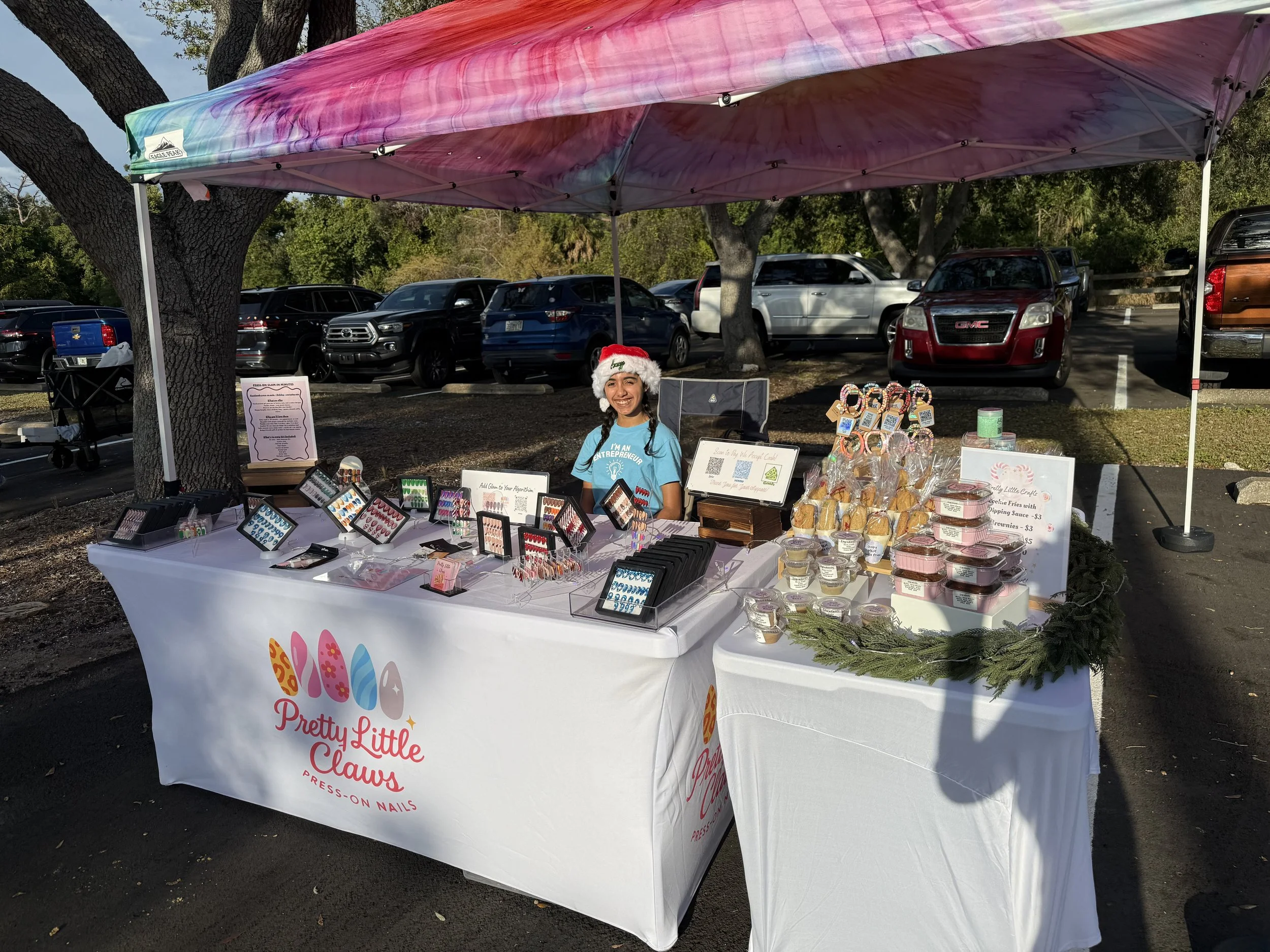 A woman smiling under a colorful pink and blue tie-dye canopy at a craft fair, standing behind a table with jewelry, baked goods, and craft items, with cars parked in the background.