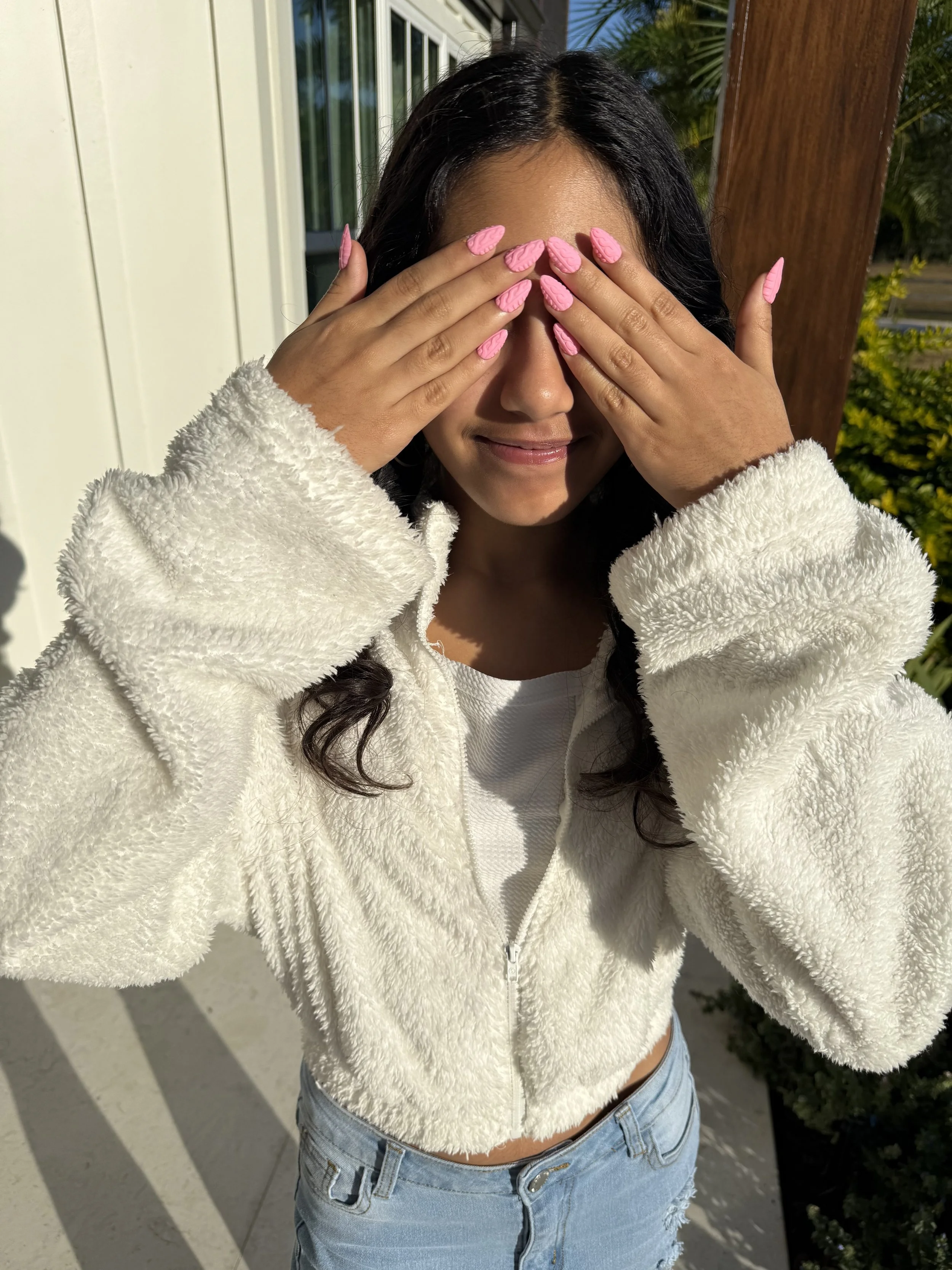 A woman with long dark hair wearing a white fleece jacket and ripped blue jeans covering her face with both hands, pink painted nails, standing outdoors with sunlight, a white door, green plants, and a wooden post in the background.