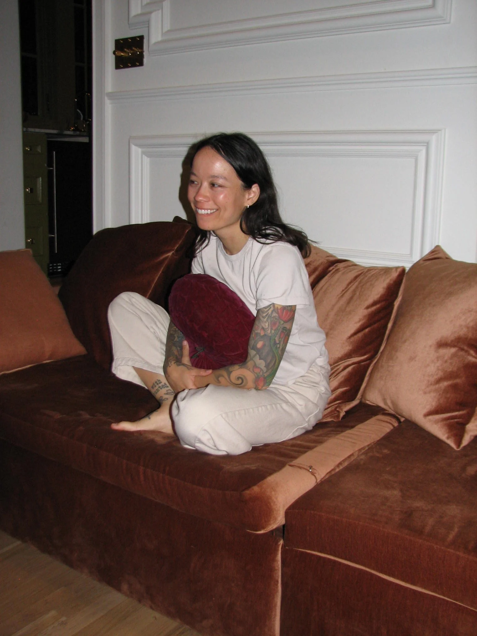Jazmin Lee is a real estate advisor in Nashville, has tattoos on her arms sitting cross-legged on a brown velvet couch, smiling and hugging a red pillow, in a room with white paneled walls.