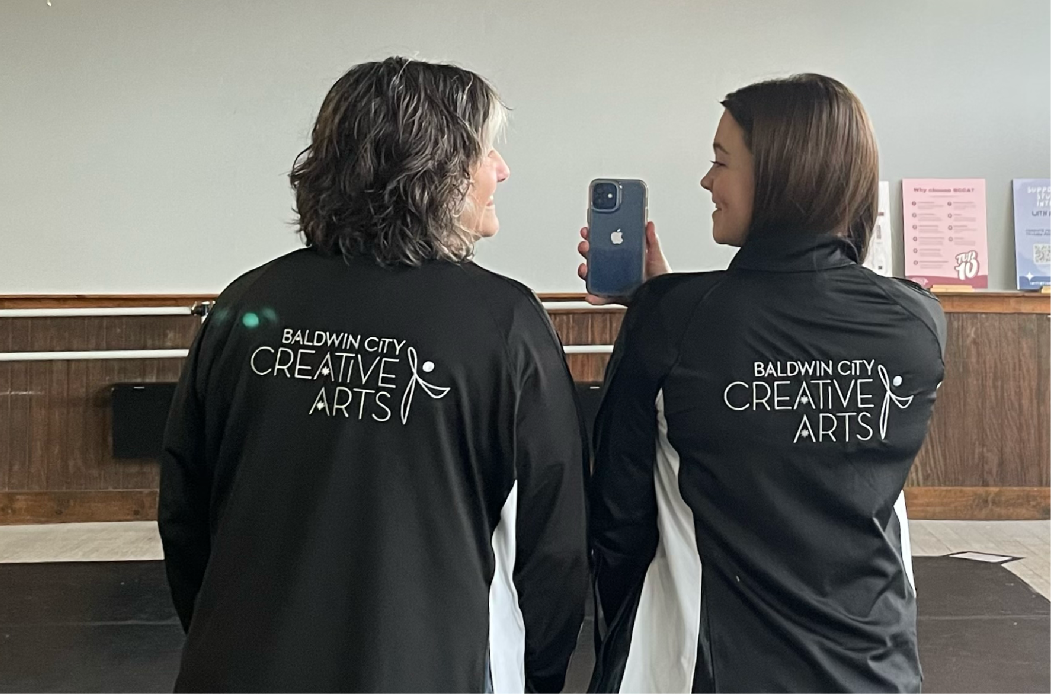 Two women are smiling and taking a photo of each other with a smartphone in an indoor setting. They are wearing black jackets with the logo 'Baldwin City Creative Arts' on the back.