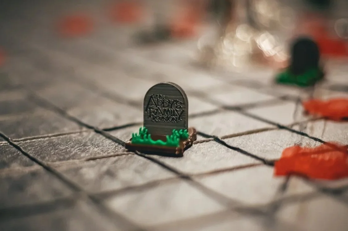 A miniature tombstone with the inscription "ADDY'S FAMILY LOVE" on a game board, surrounded by tiny orange leaves.