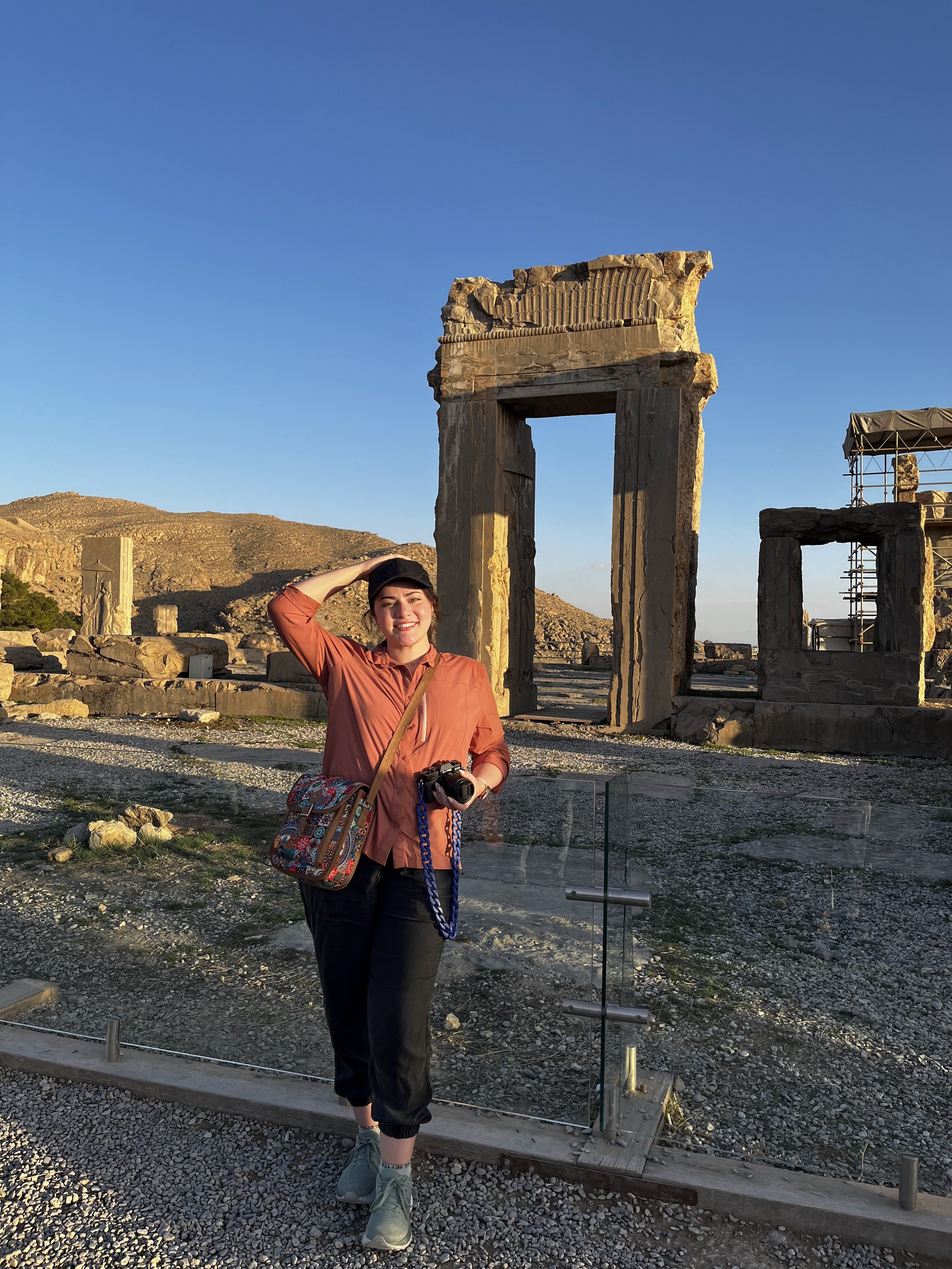 A woman standing at an ancient archaeological site, with stone ruins and columns around her, smiling and holding a camera, during sunset or late afternoon.