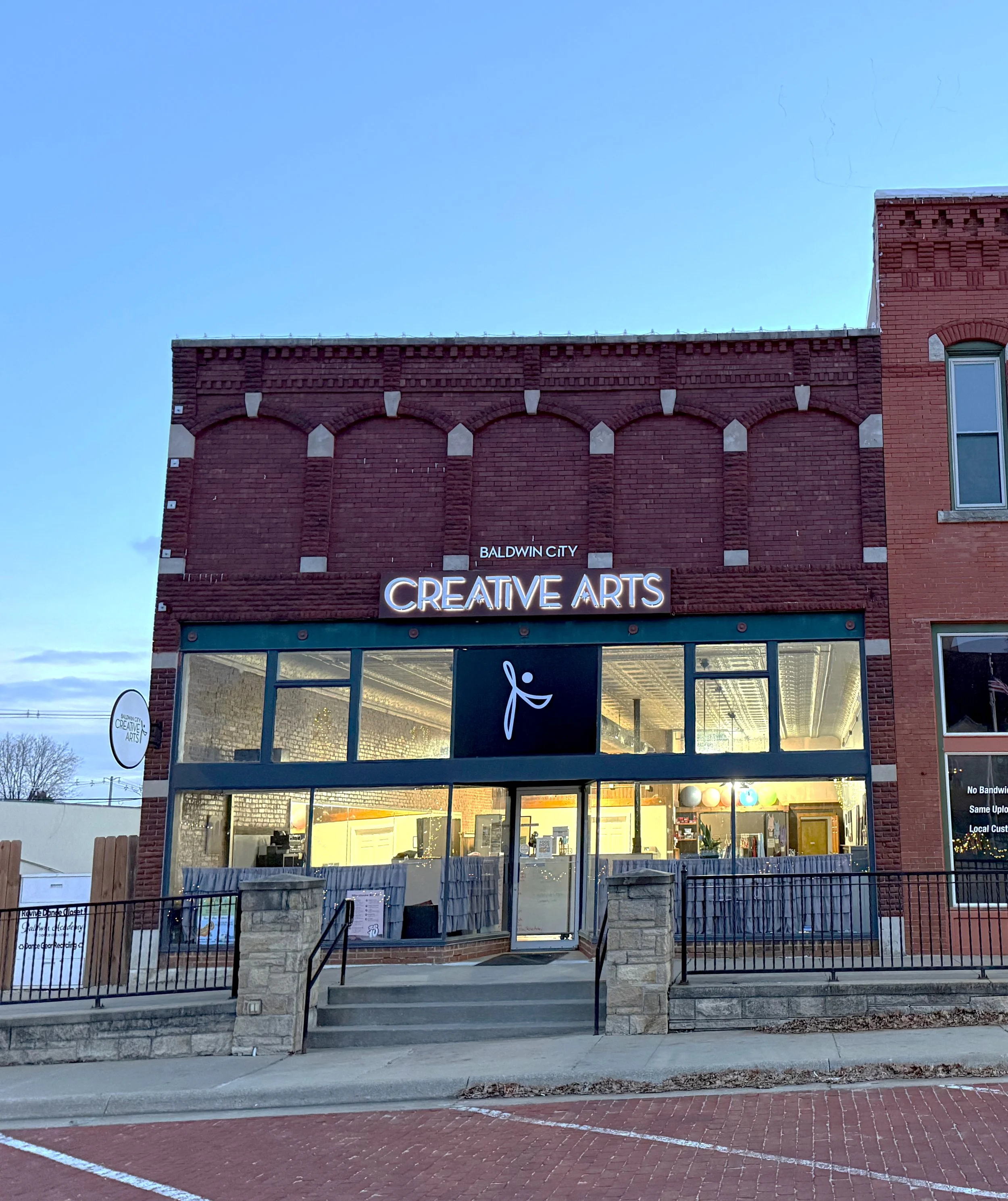 Front view of a brick building with a signage that reads 'Baldwin City Creative Arts' in neon lights, large glass windows on the ground floor, and a small circular sign on the side also displaying 'Baldwin City Creative Arts.' The building has a dark blue door and is situated in a brick-paved parking area with a sidewalk and a black metal fence in front.