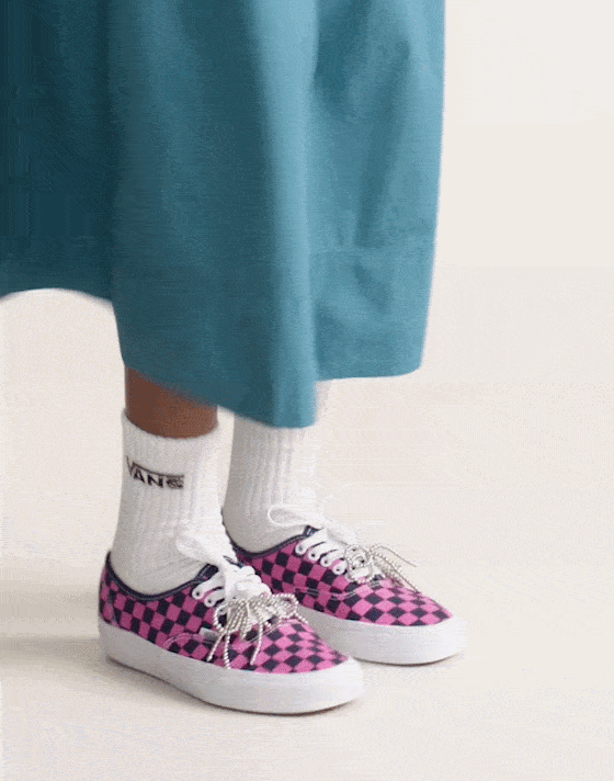 Close-up of a person's feet wearing pink and black checkered sneakers, white socks with black logo, and teal skirt.