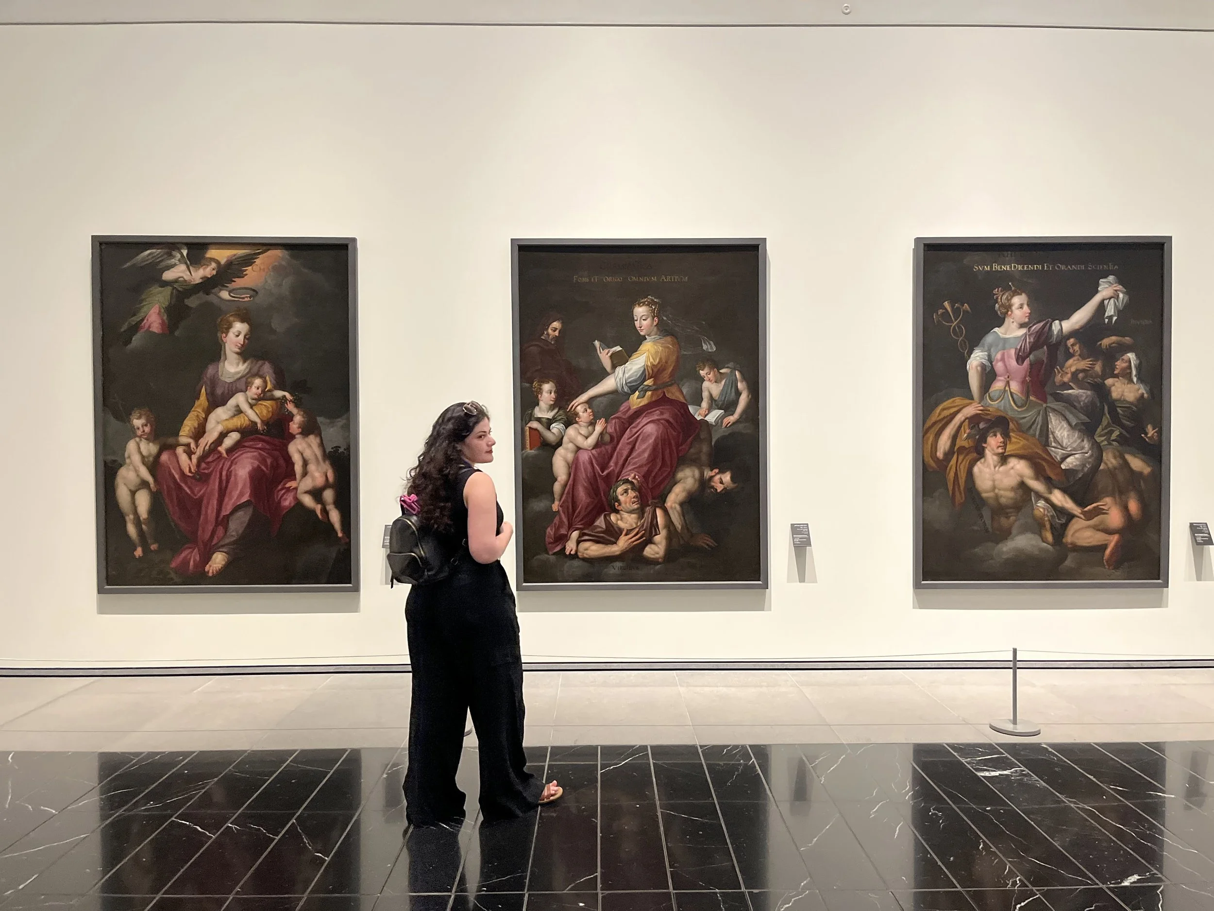 A woman with dark, wavy hair in a black top standing in front of three large classical paintings in a museum.