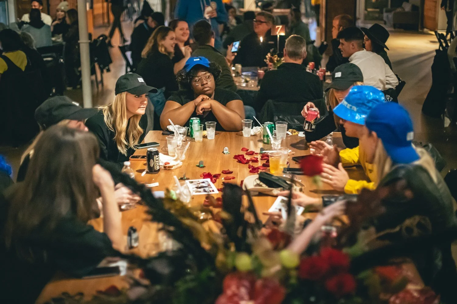 People gathered around a long wooden table at a social event, some wearing hats or casual clothing. The table has scattered rose petals, drinks, and personal items, with a lively background of more groups and decorations, suggesting an indoor or outdoor party atmosphere.