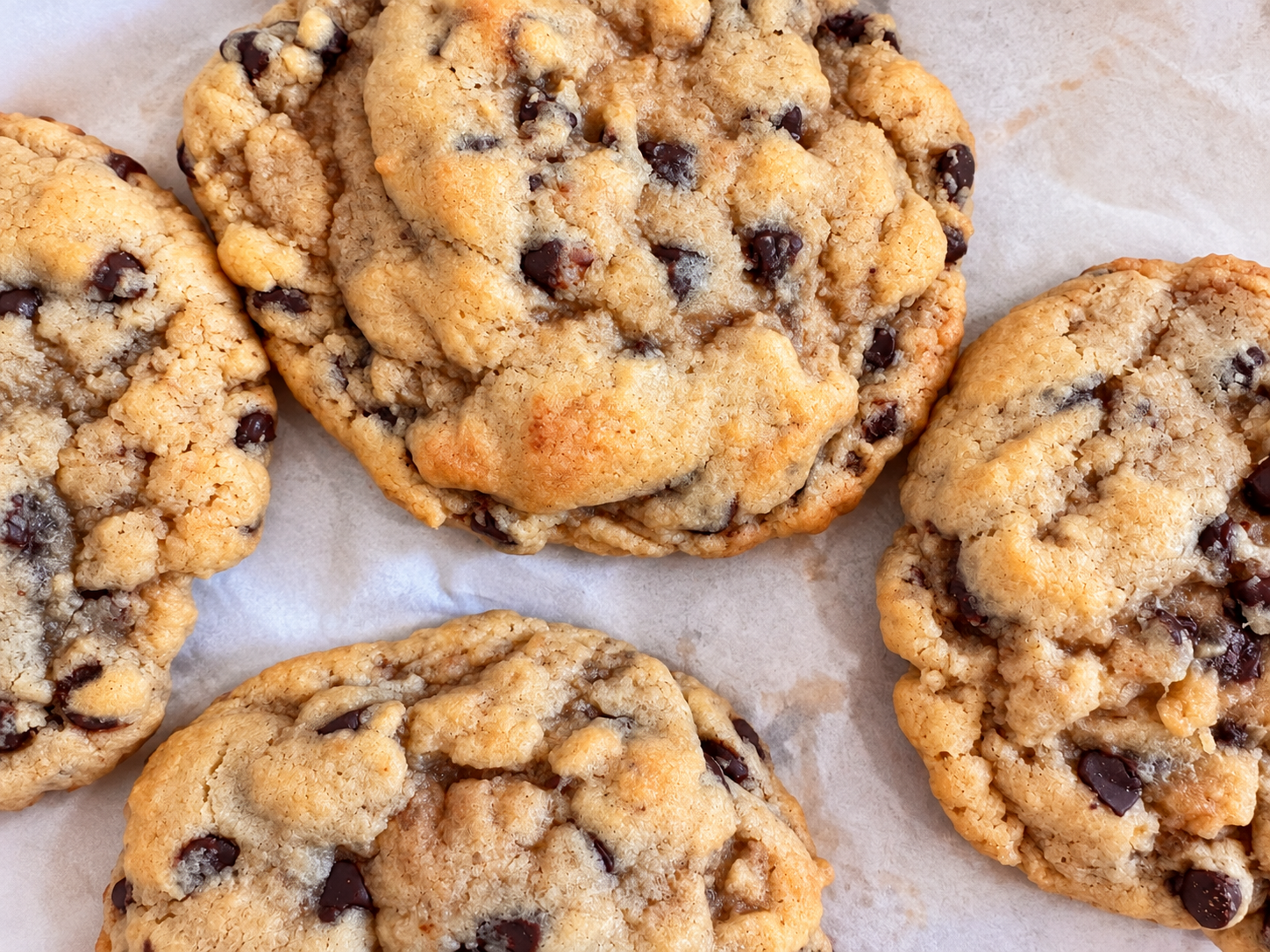 Close-up of five chocolate chip cookies on parchment paper, showing their golden-brown crispy edges and soft centers with dark chocolate chips.
