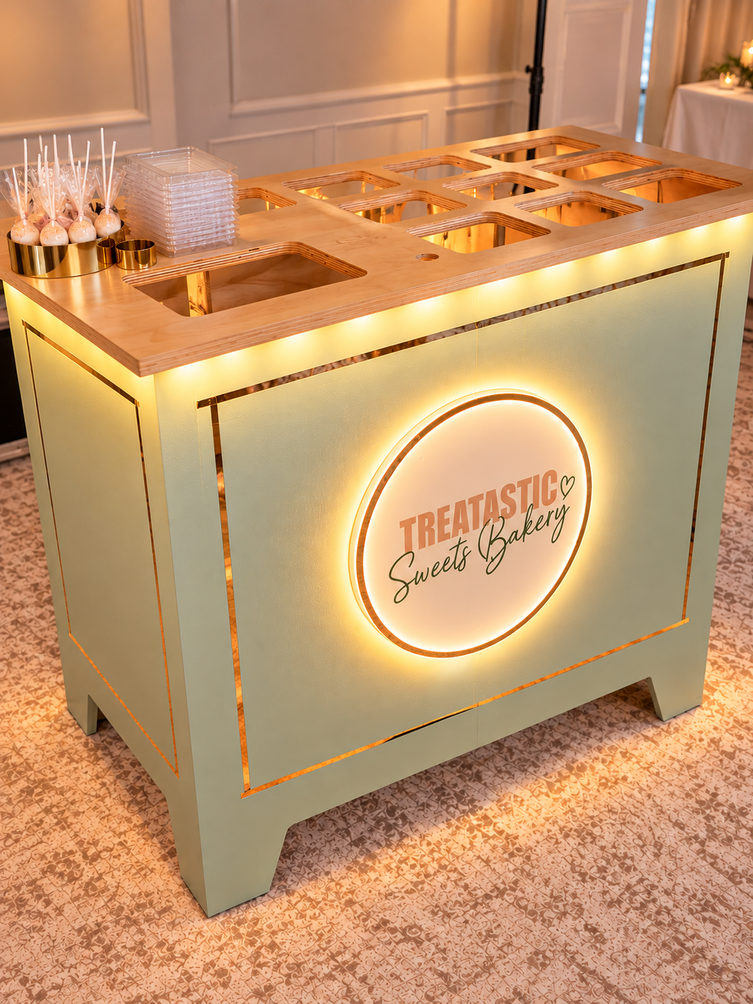 A dessert stand with the sign 'Treatastic Sweets Bakery' illuminated with LED lights. The stand has empty compartments for treats, with some skewered treats and plastic containers on top.