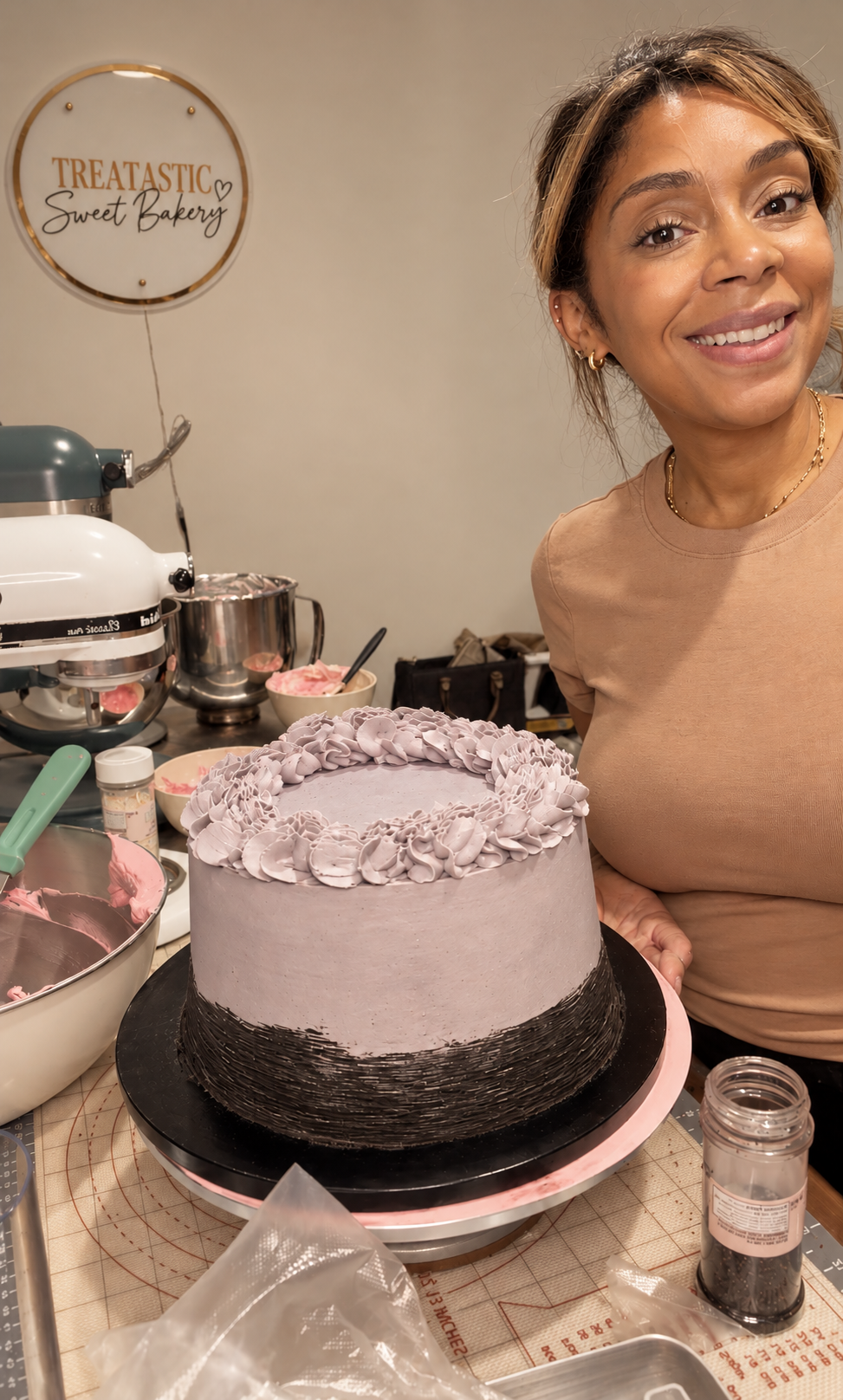 A woman is smiling next to a decorated cake on a party prep table. The cake has purple frosting with darker coloring at the bottom and purple floral decorations on top. The background includes baking tools and bowls, and a sign that reads 'TREATASTIC Sweet Bakery' on the wall.