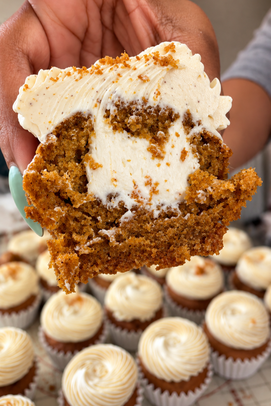 A person holding a large slice of carrot cake with cream cheese frosting, with cupcakes decorated with swirled white frosting in the background.