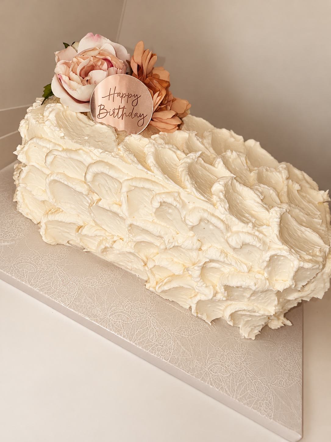 Cream-colored birthday cake with floral decorations and a 'Happy Birthday' topper on a white textured cake board.