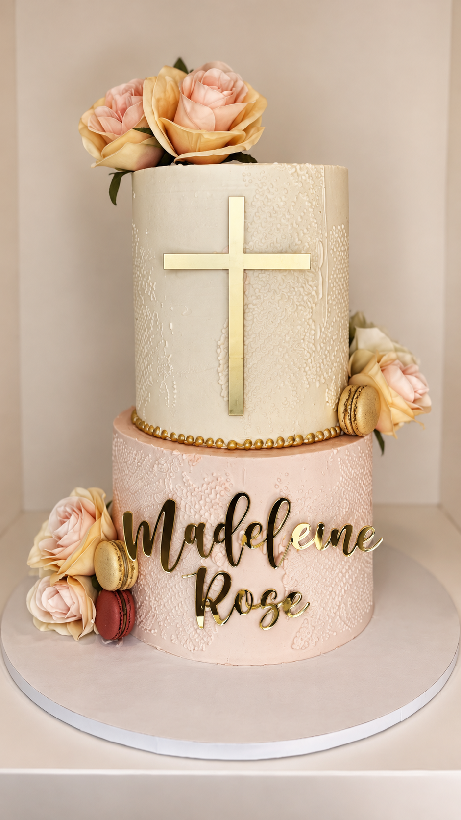 A two-tiered cake decorated with pink roses, macarons, and a gold cross, with a gold "Madeleine Rose" sign on the lower tier.