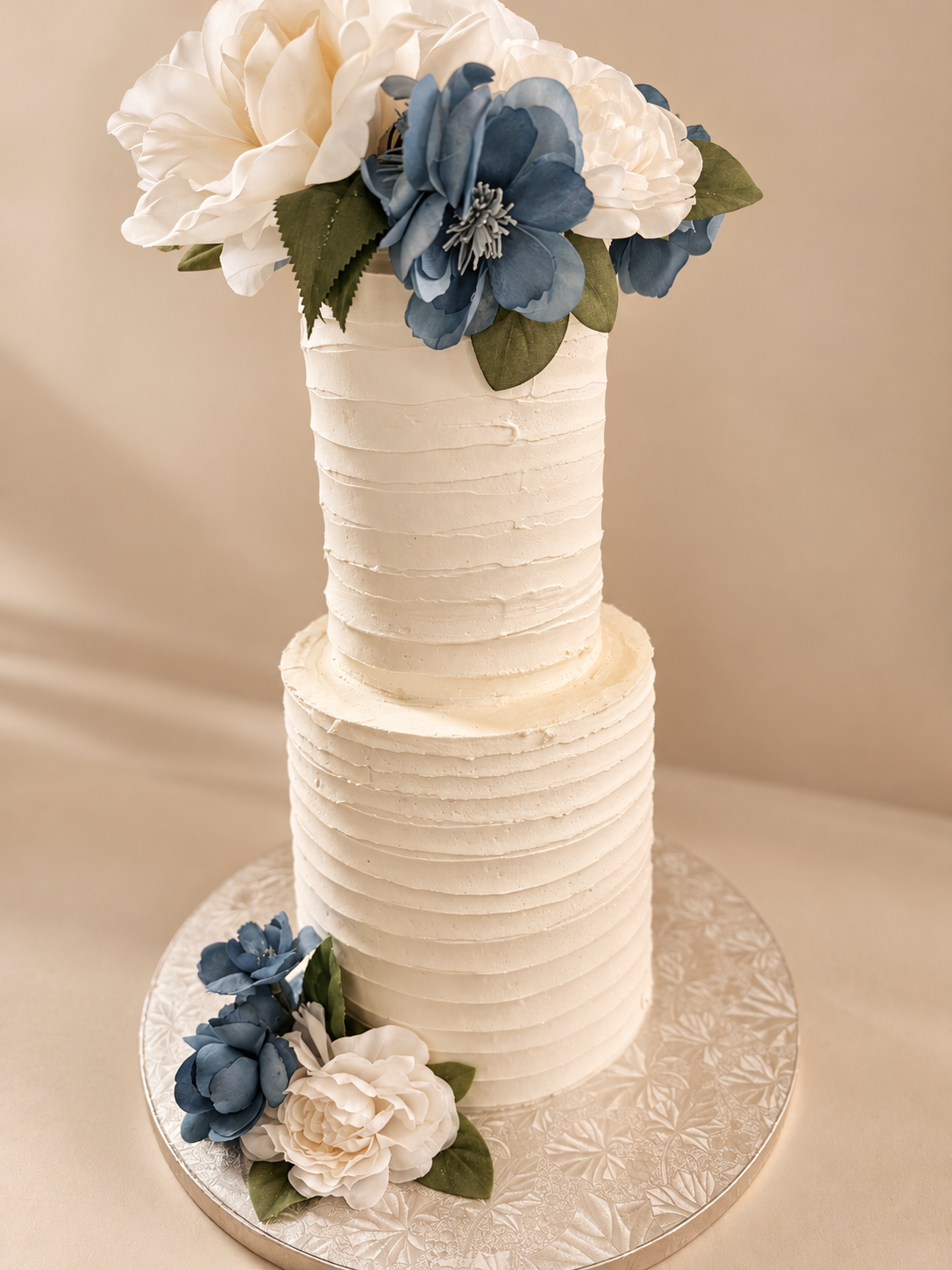 A two-tiered white wedding cake with a textured, ridged frosting design, decorated with white and blue artificial flowers, and placed on a silver, patterned cake board.