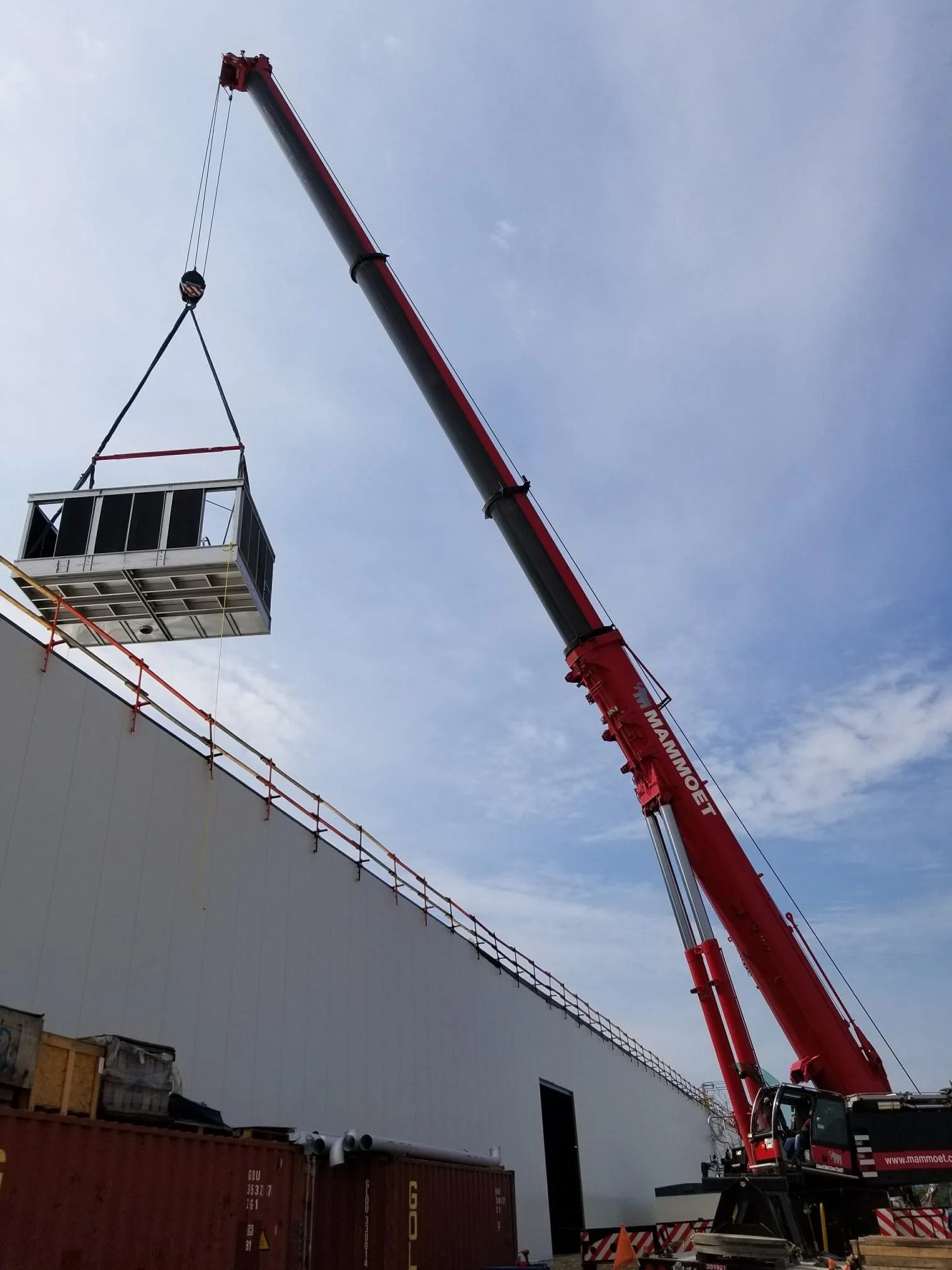 A large red crane lifting a white container with black panels at a construction site. The crane is positioned next to a tall gray wall with a blue sky in the background.