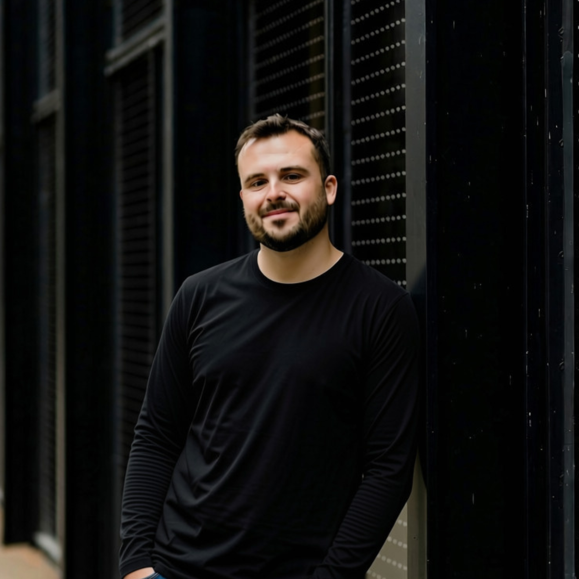 Young man with short dark hair and beard smiling, wearing a black long-sleeved shirt, standing in front of a black metal fence with vertical slats.
