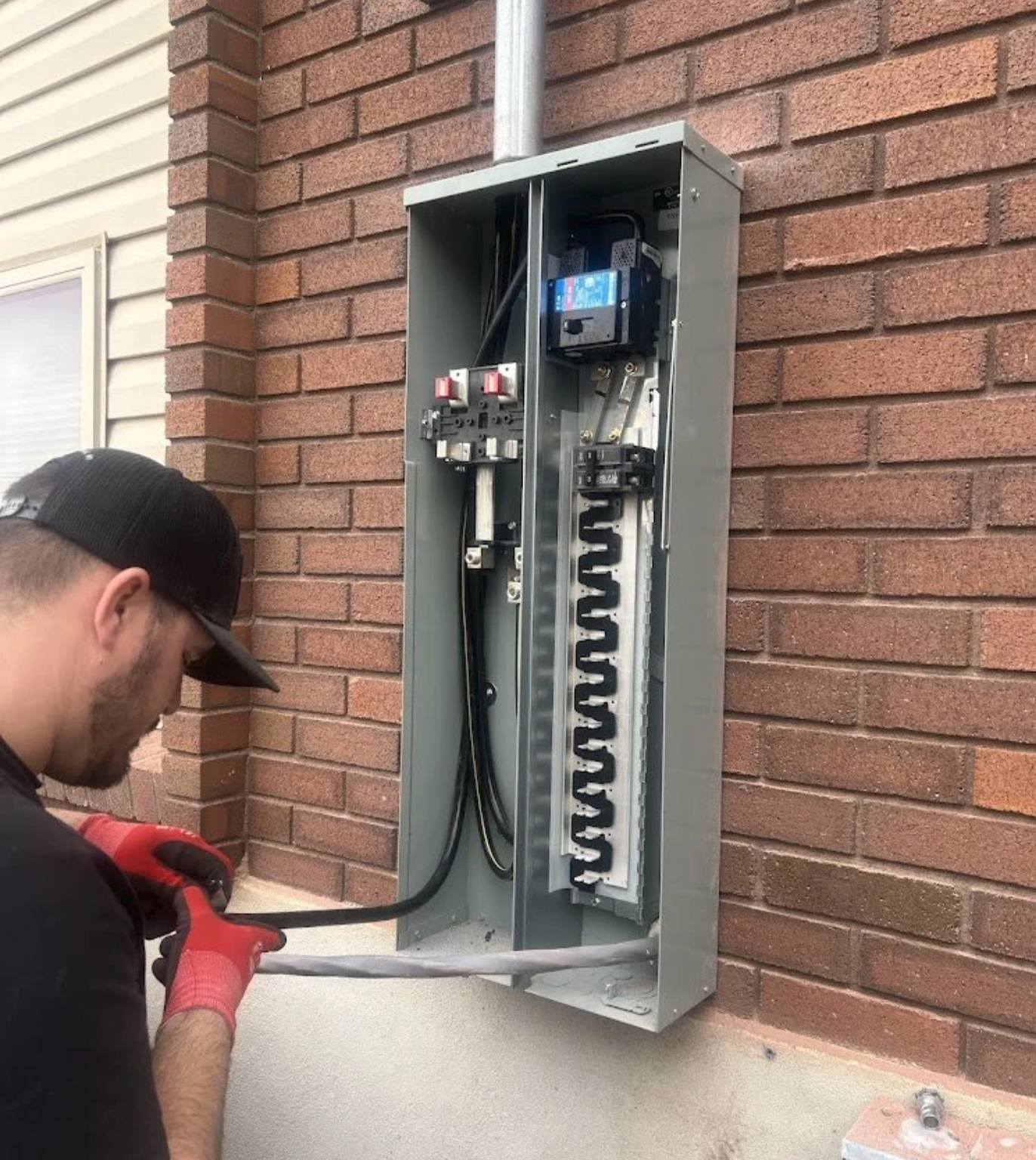 A technician installing or repairing an outdoor electrical panel box on a brick wall, with a screwdriver in hand and tools on his belt.