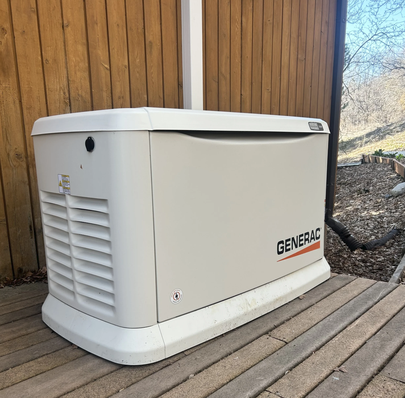 A Generac green energy generator on a wooden deck with a wooden fence and trees in the background.