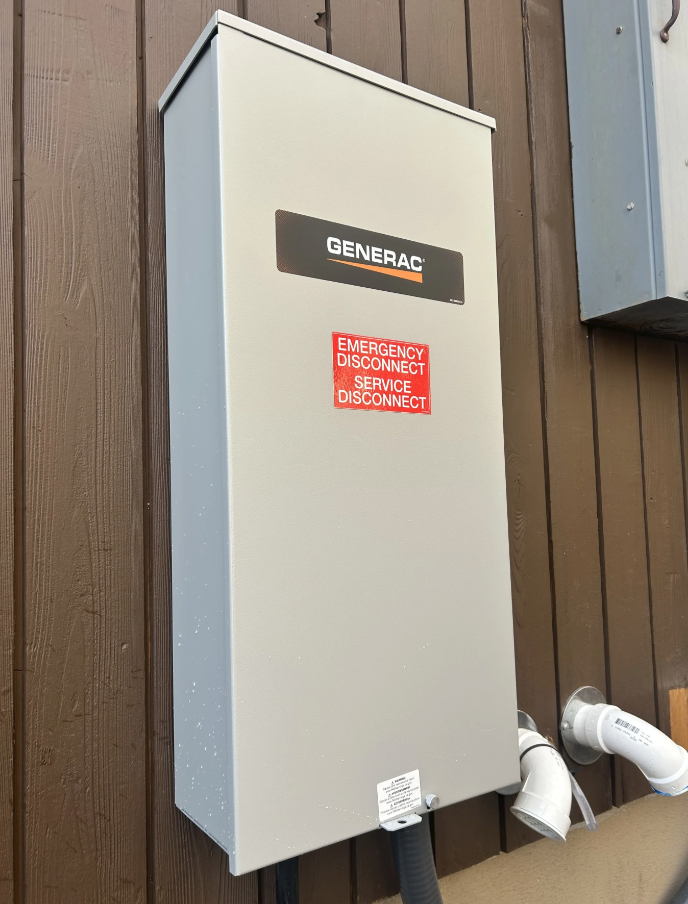 Electrical generator panel mounted on a brown wooden wall with warning label and connected pipes.