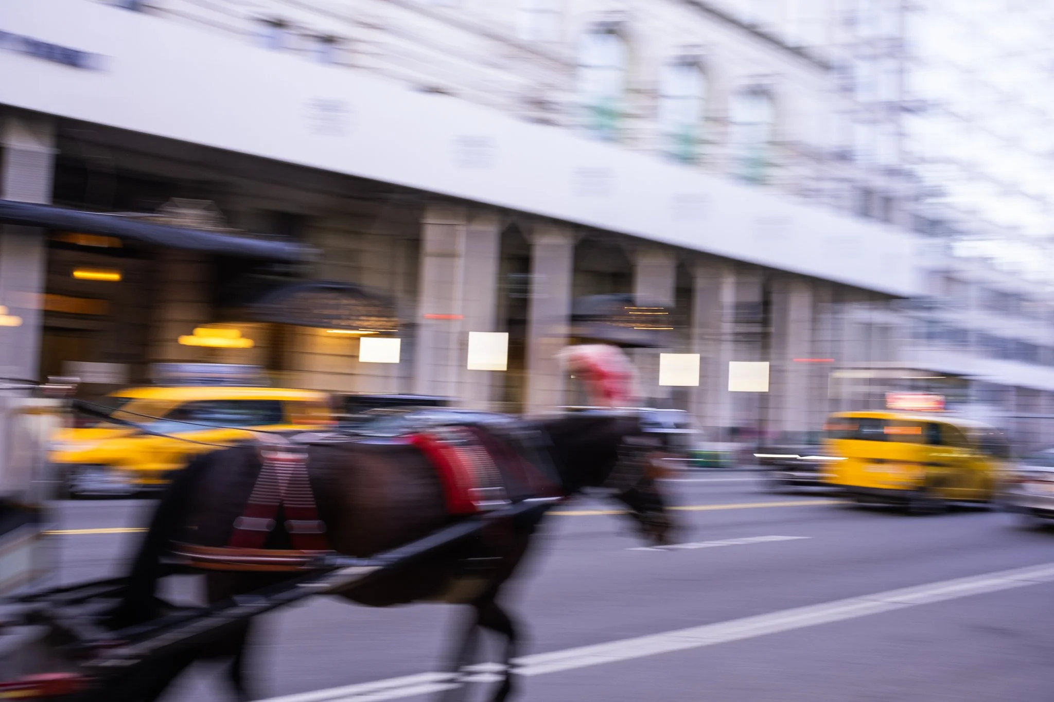 Blurred city street scene with yellow taxis, parked cars, and building with storefronts.