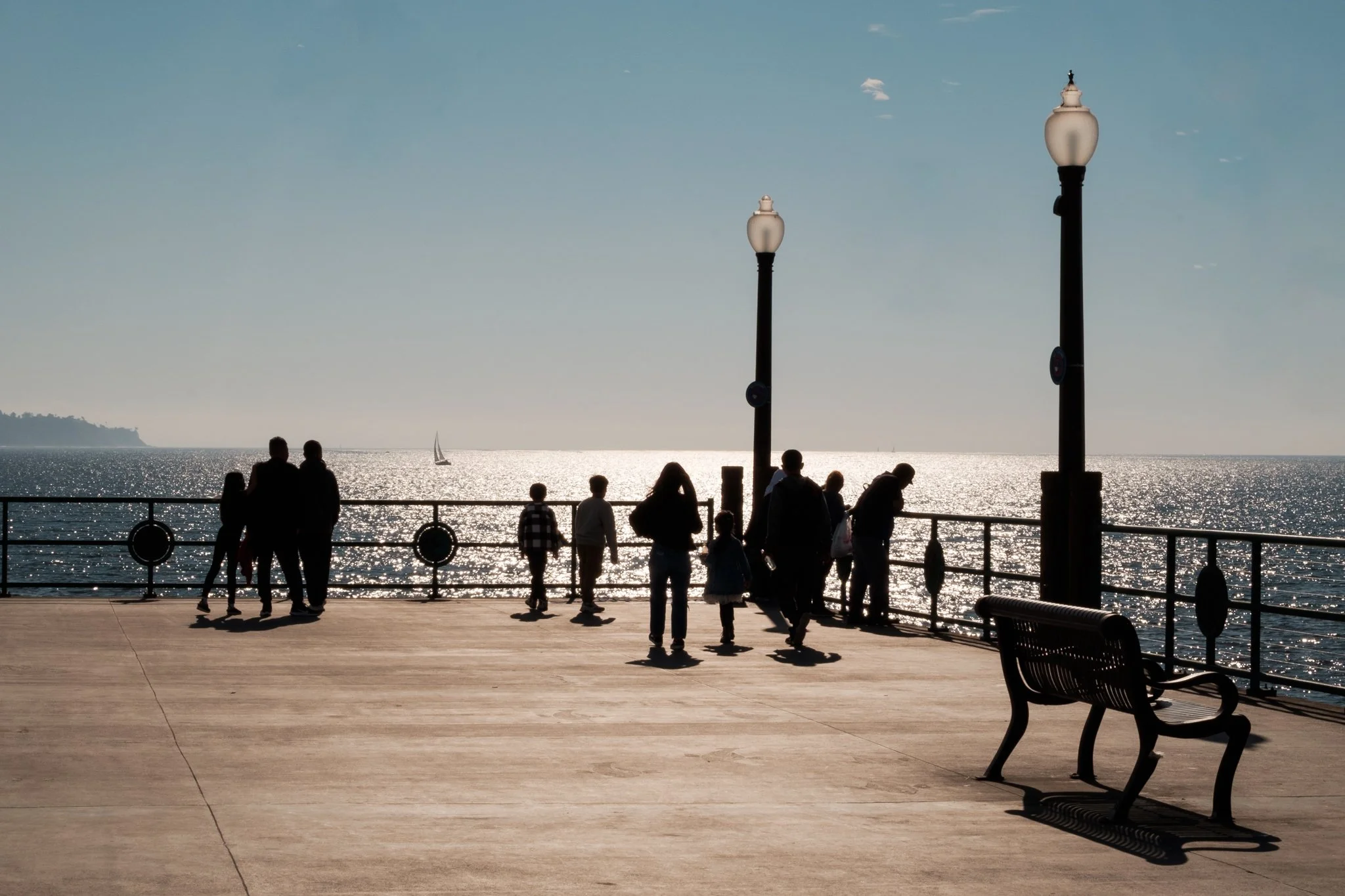 People walking and sitting along a waterfront promenade at sunset with a sailboat in the water and lampposts lining the walkway.