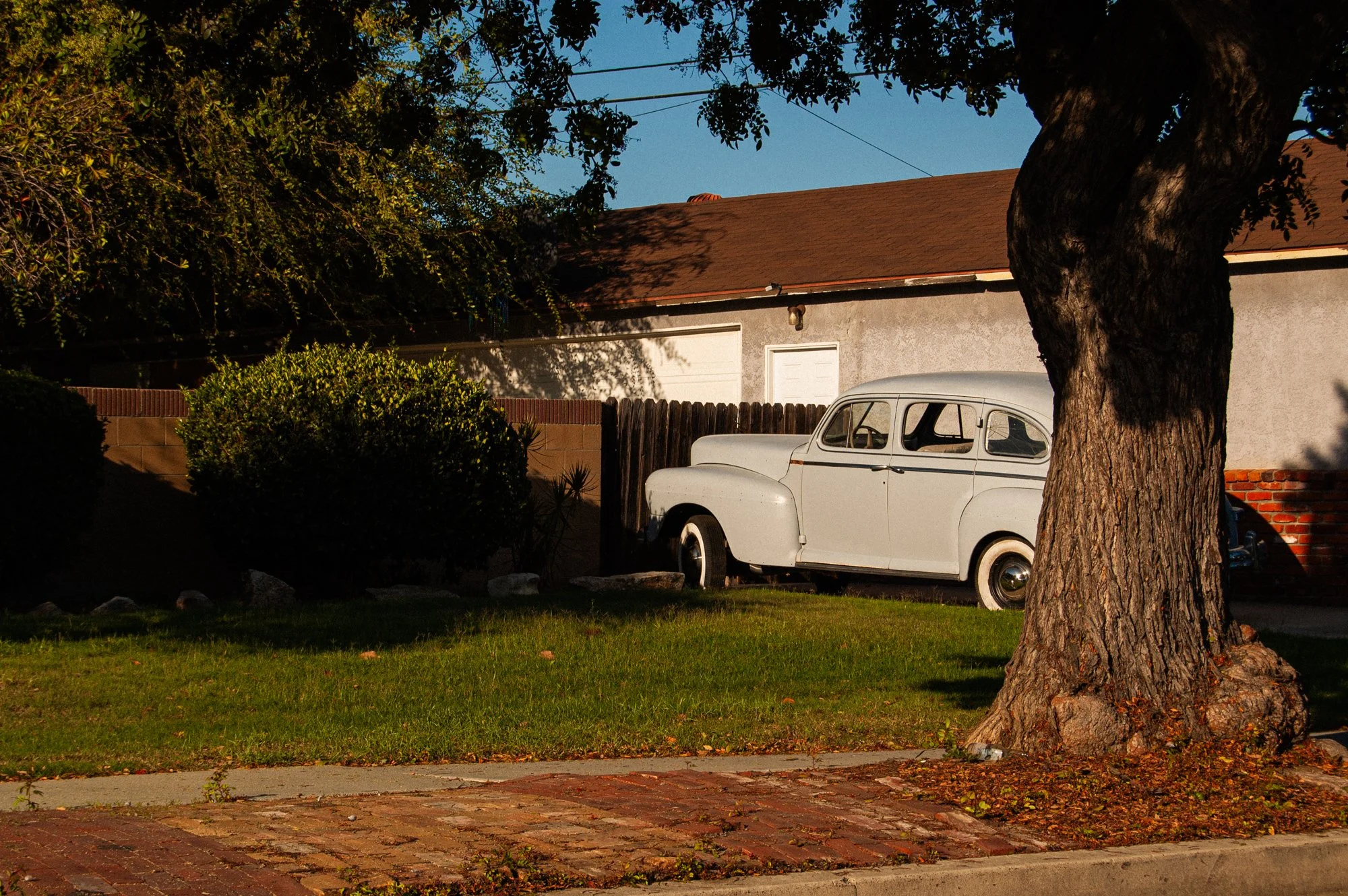 A vintage white car parked under a large tree on a suburban street. The house behind has a red shingle roof and a gray stucco exterior.