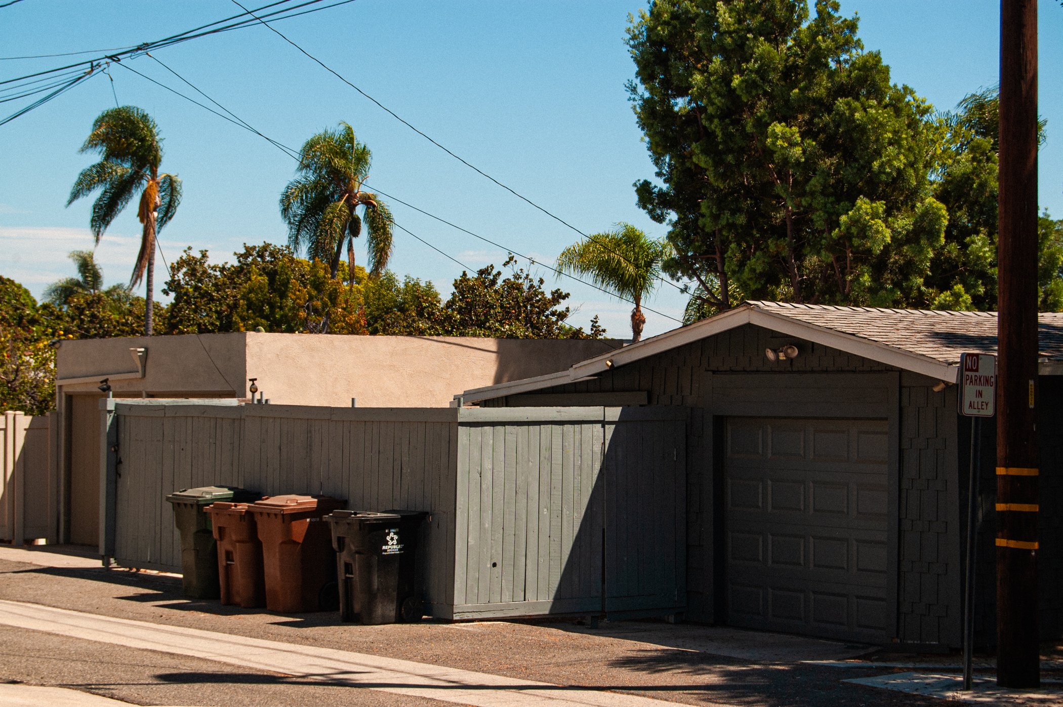 Backyard alley with trash bins, a gray wooden fence, and a garage. Tall trees and palm trees under a clear blue sky. Electric wires overhead.
