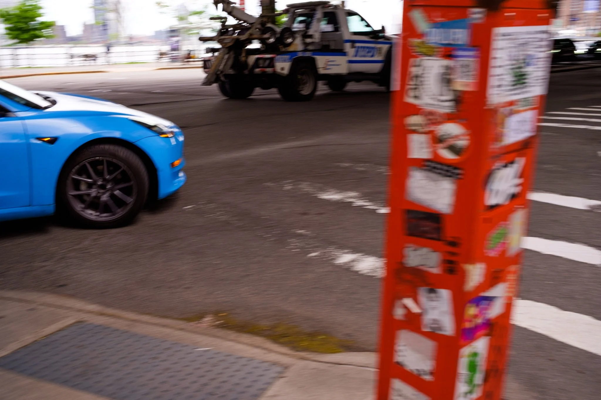 Blurred street scene with a blue sports car, a tow truck, and a red vending machine or advertisement stand in the foreground.