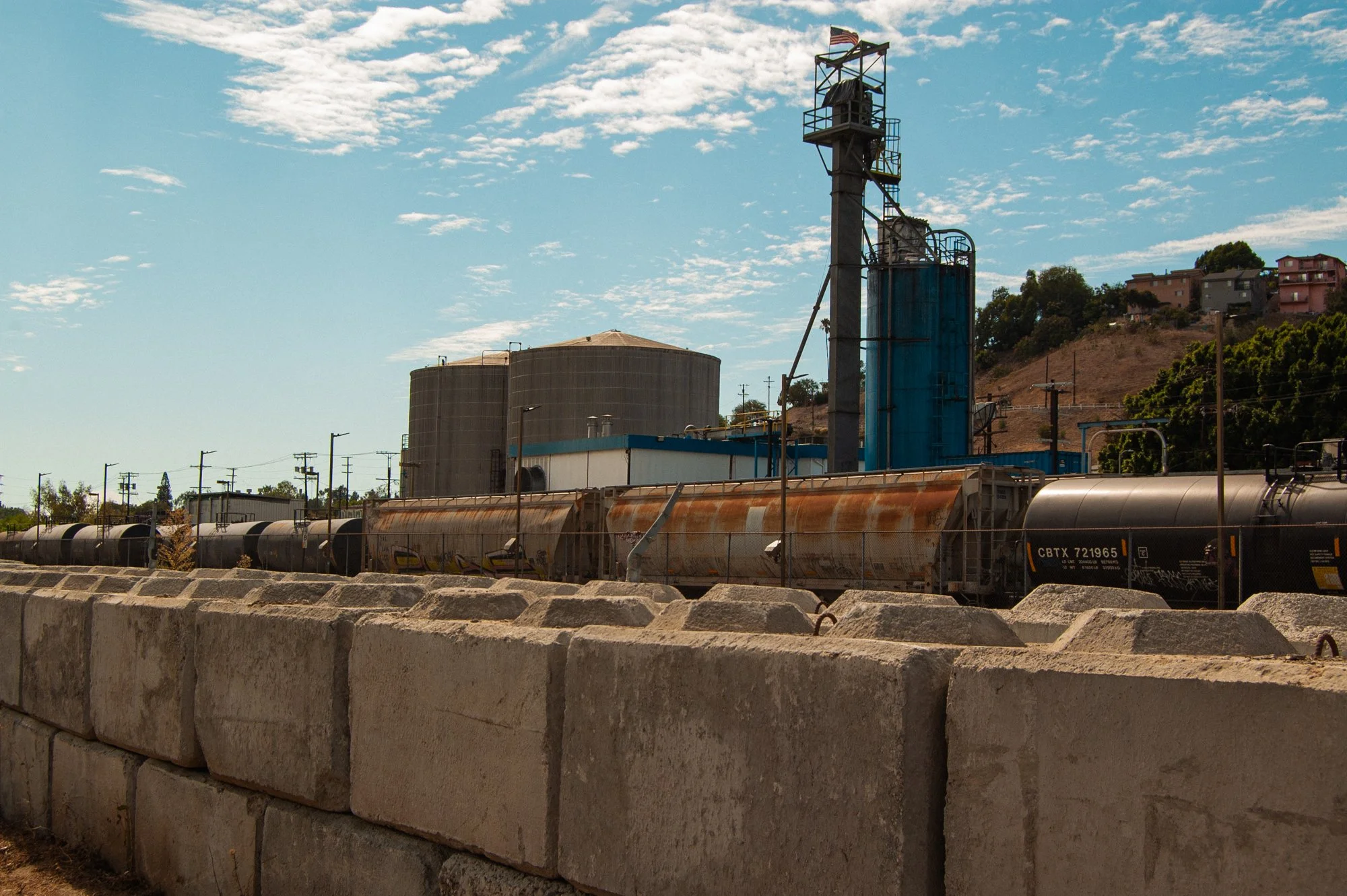 A row of concrete blocks forming a low wall in the foreground, a freight train with tanker cars behind it, and industrial buildings with silos and a tall structure with an American flag on top, set against a hillside with houses and trees under a par