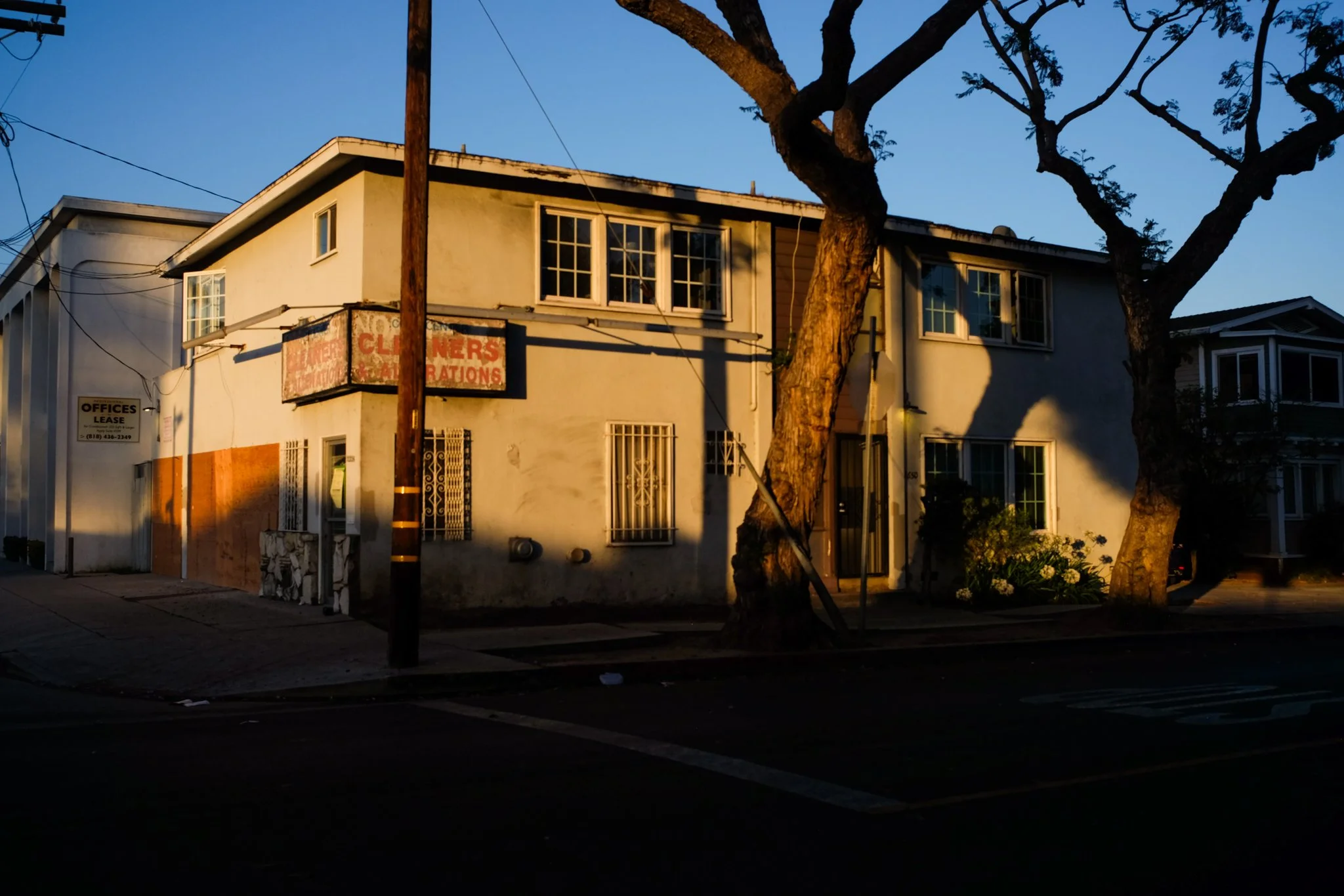 A two-story beige building with multiple windows, some barred, and a faded sign near the top that reads 'NO CLINER' and other text. A small sign on the side indicates office space for lease. A large tree with twisted branches is in front, and the bui