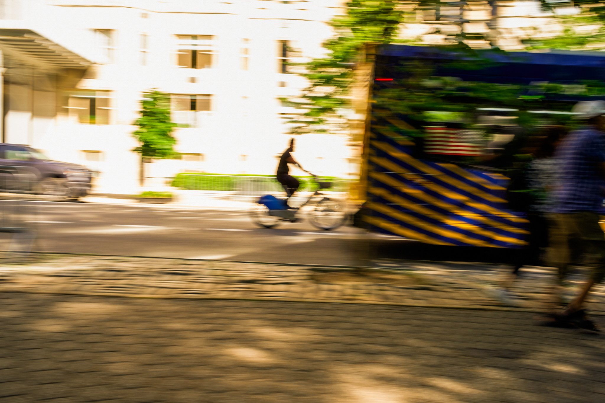 A person riding a bicycle on a city street, with a large truck partially blocking the view, and buildings in the background.