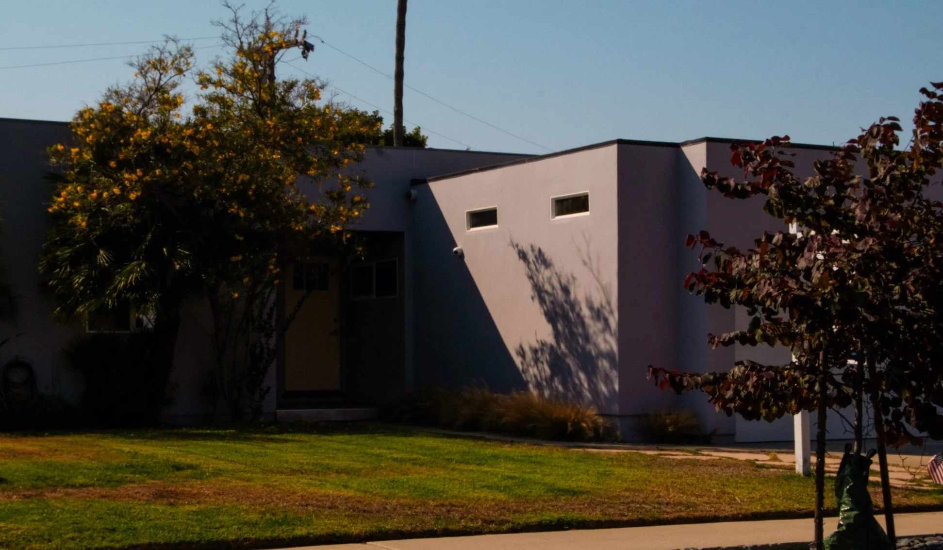 A modern white residential building with small rectangular windows, surrounded by trees with autumn-colored leaves, and a well-maintained grass lawn.