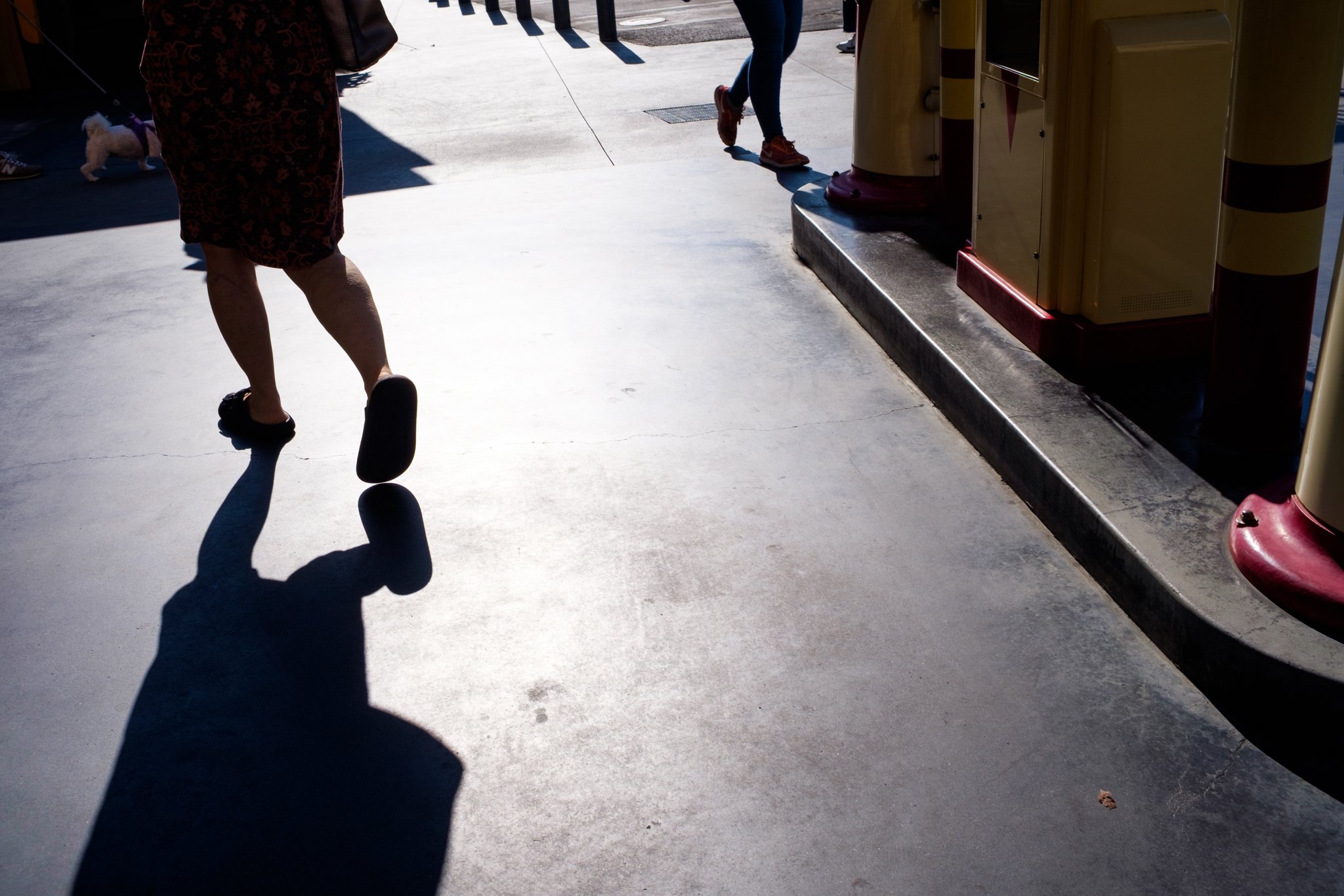 Part of a sidewalk and a drop-off with a yellow and red parking barrier arm on the right. Several people are walking, and the shadows suggest it is daytime with a sunny sky.