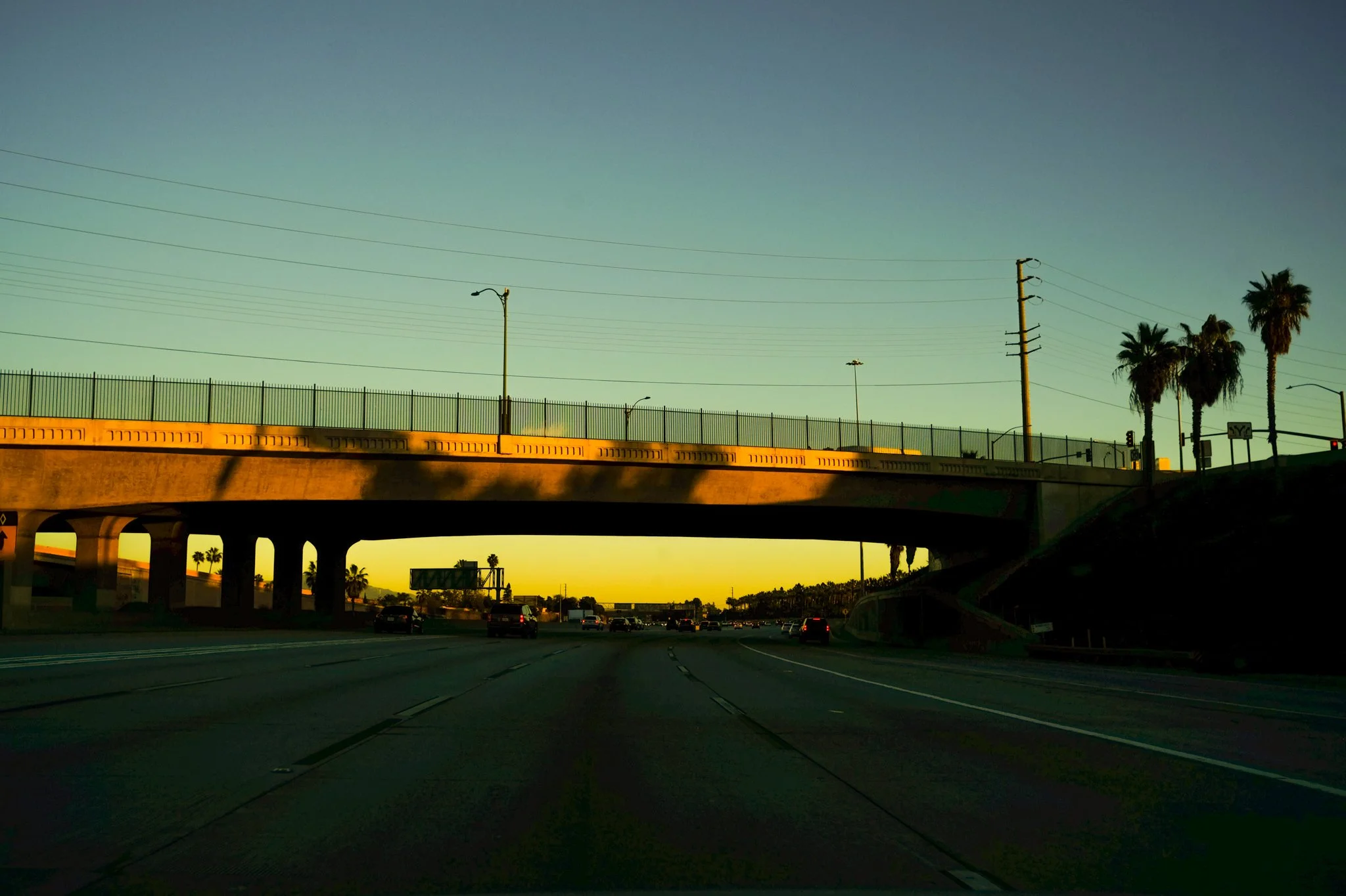 Sunset view of a highway with cars, palm trees, electric poles, and streetlights under a clear sky.
