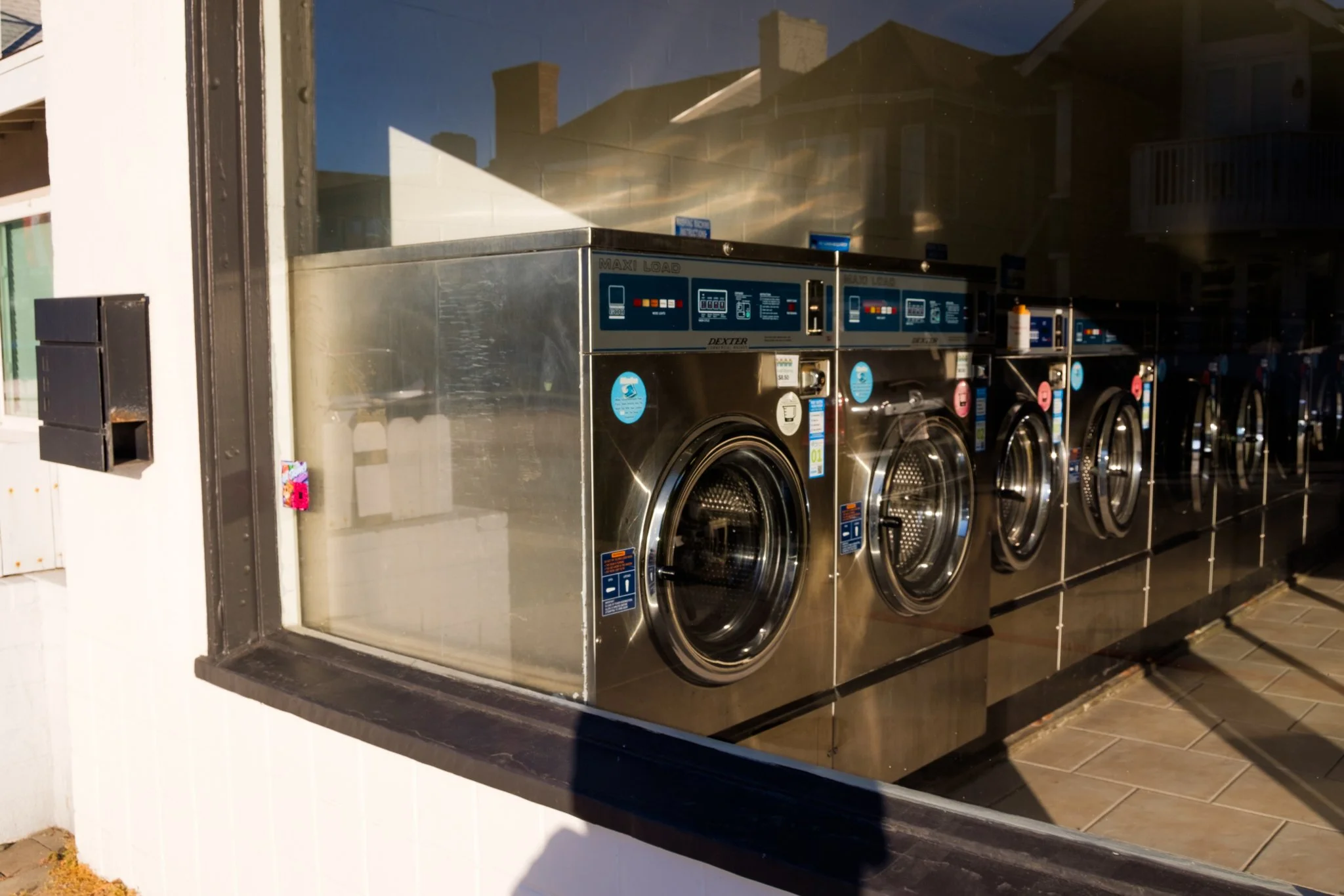 A row of stainless steel commercial washing machines with digital controls and round glass doors, positioned against a reflective glass window showing a house and blue sky.
