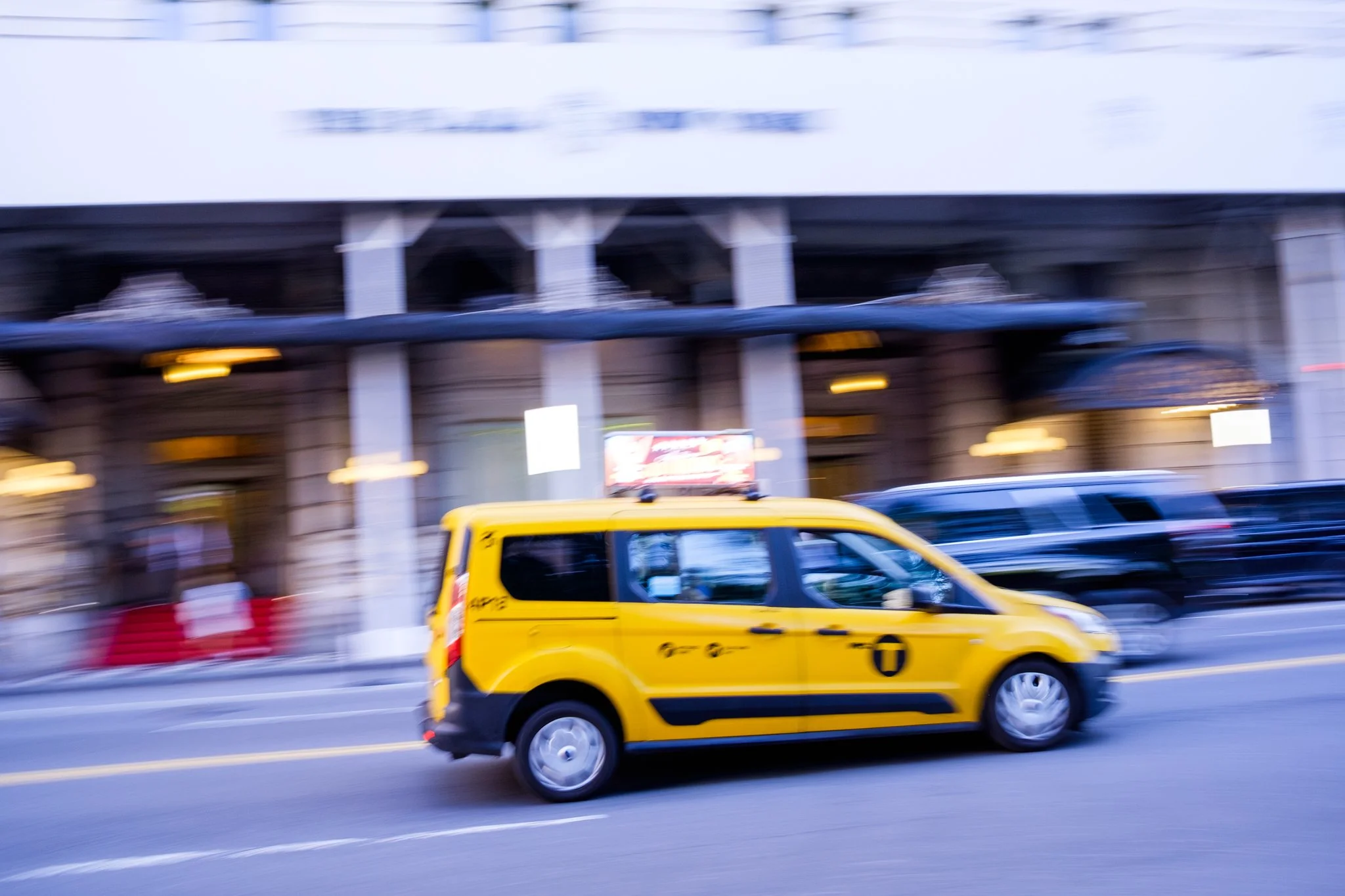 Blurred city street scene showing a moving yellow taxi cab and other vehicles passing under an elevated train or bridge.