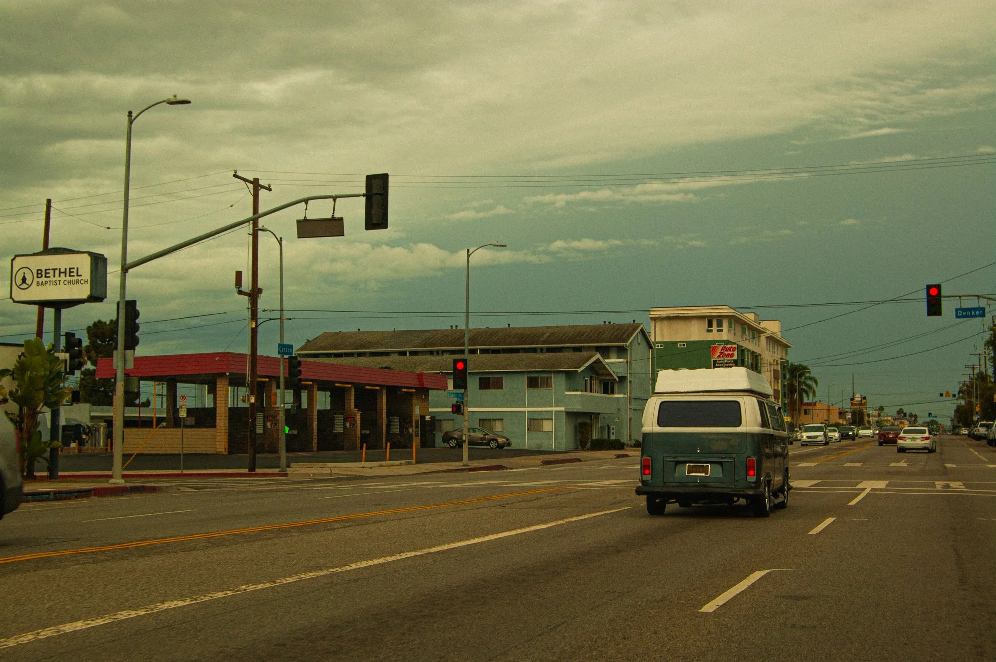An urban street scene with a blue and white van stopped at a red traffic light, cars ahead, and buildings including a Methodist church and a gas station in the background, under cloudy skies.
