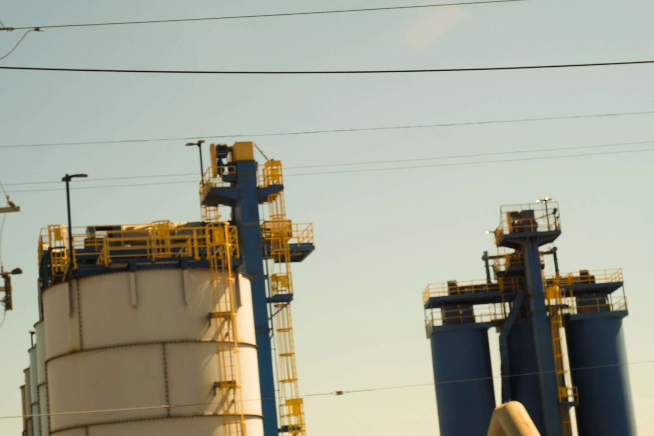 Industrial facility with large white tanks, yellow railings, and blue structures under a pale sky with power lines.