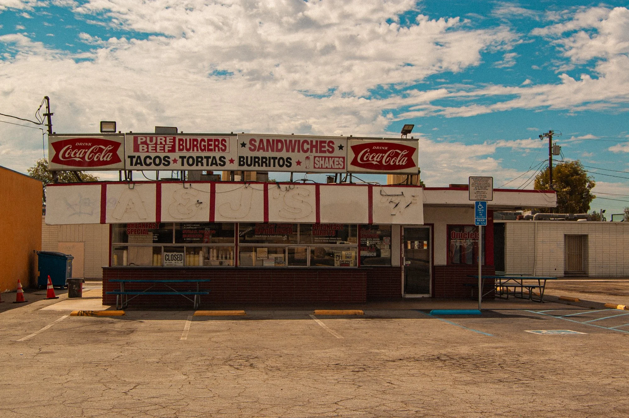 Empty fast food stand with signs advertising burgers, sandwiches, tacos, tortas, burritos, and shakes. The stand has Coca-Cola logos on top and is located in a nearly empty parking lot under a partly cloudy sky.