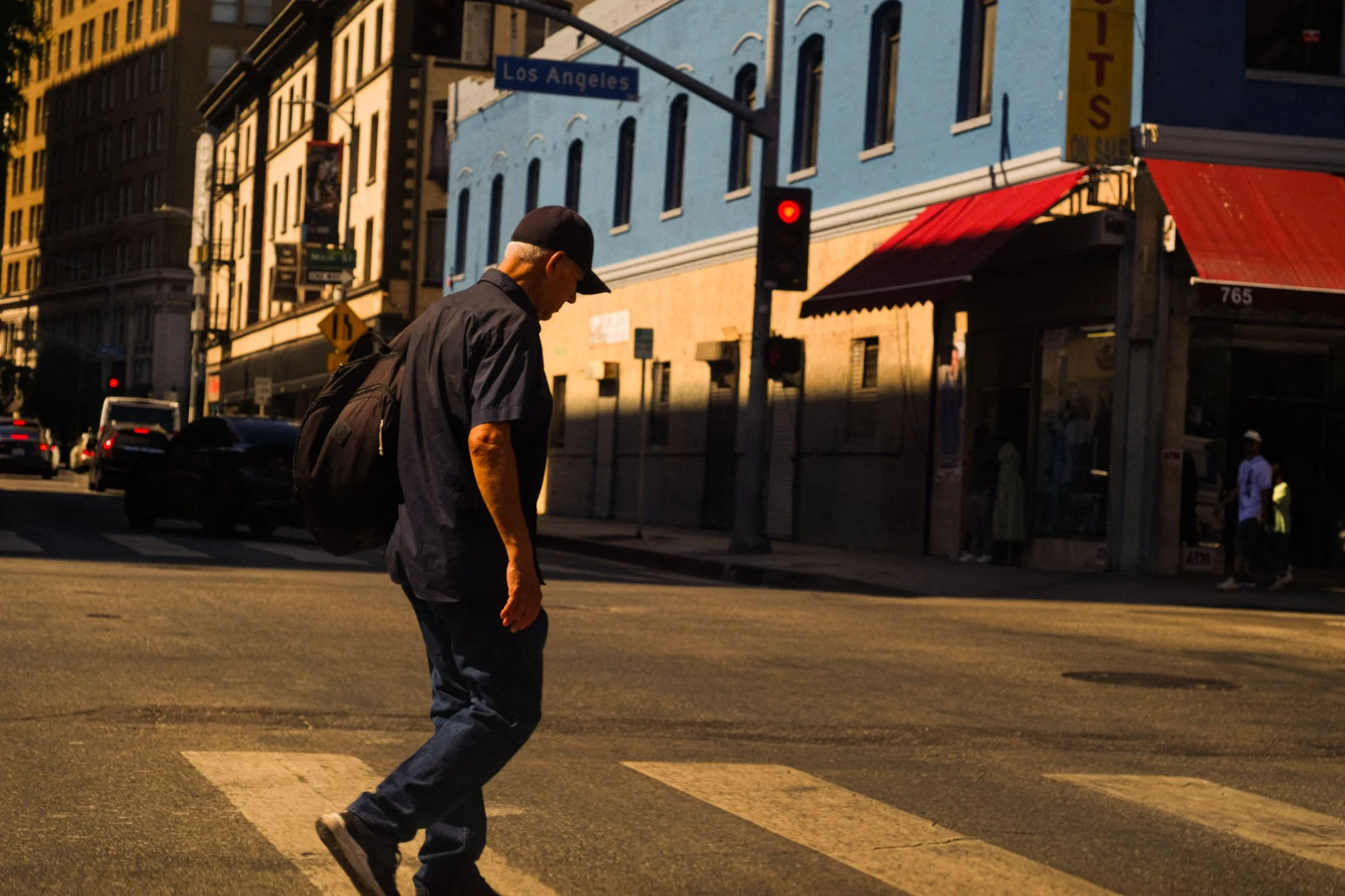 A person with a backpack crossing the street at a crosswalk in Los Angeles during sunset or dusk. There are buildings and cars visible in the background.