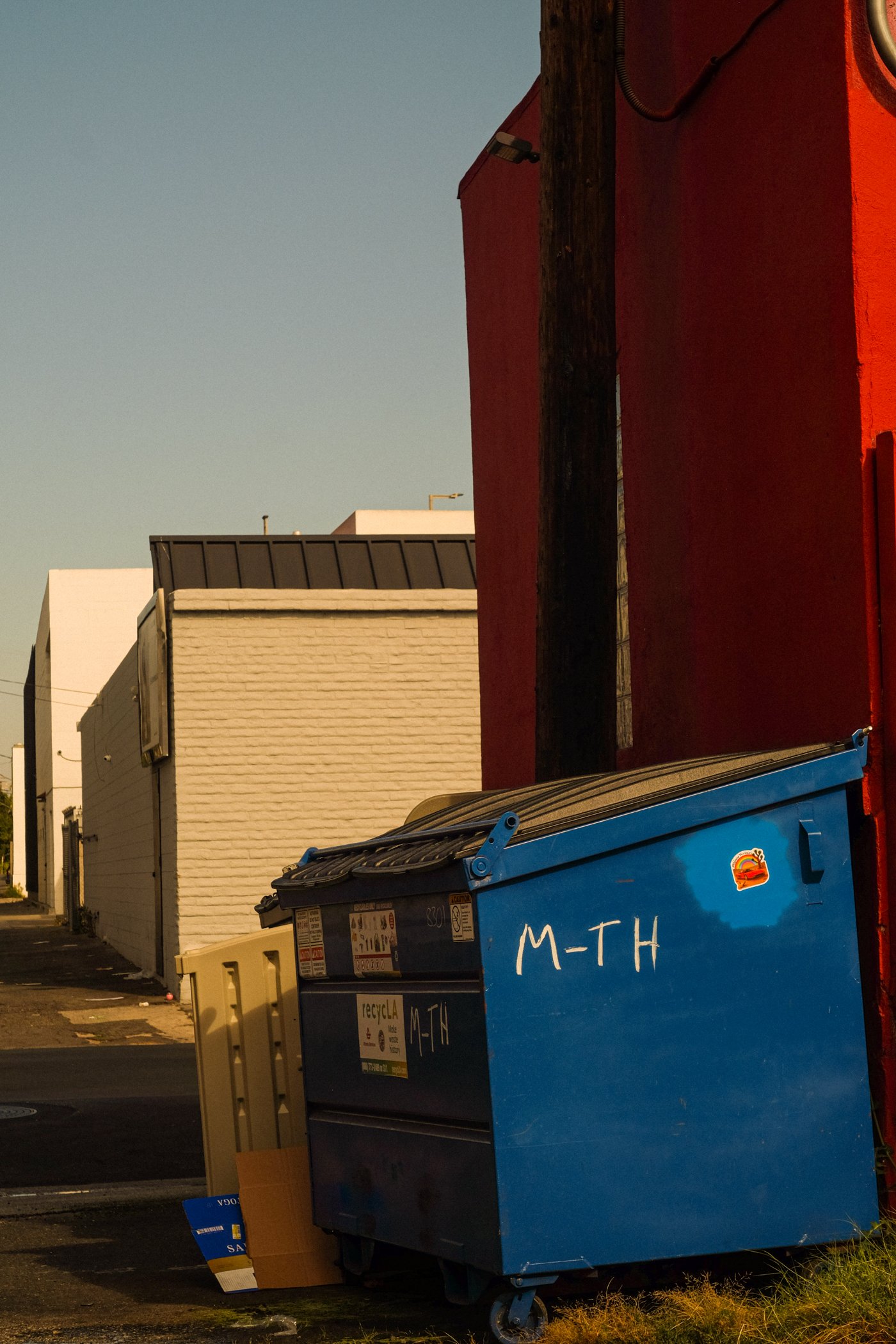 A photo of a blue recycling bin and a yellow trash bin on the street with building facades behind them, including a red and white building, under a clear sky.