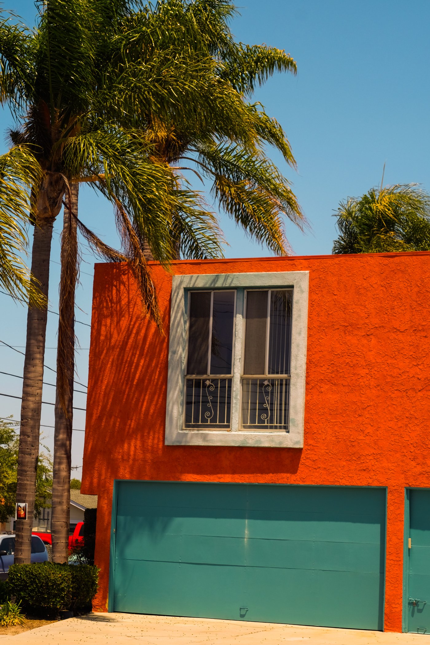 A vibrant orange building with a large teal garage door and a window with security bars above it, flanked by palm trees under a clear blue sky.