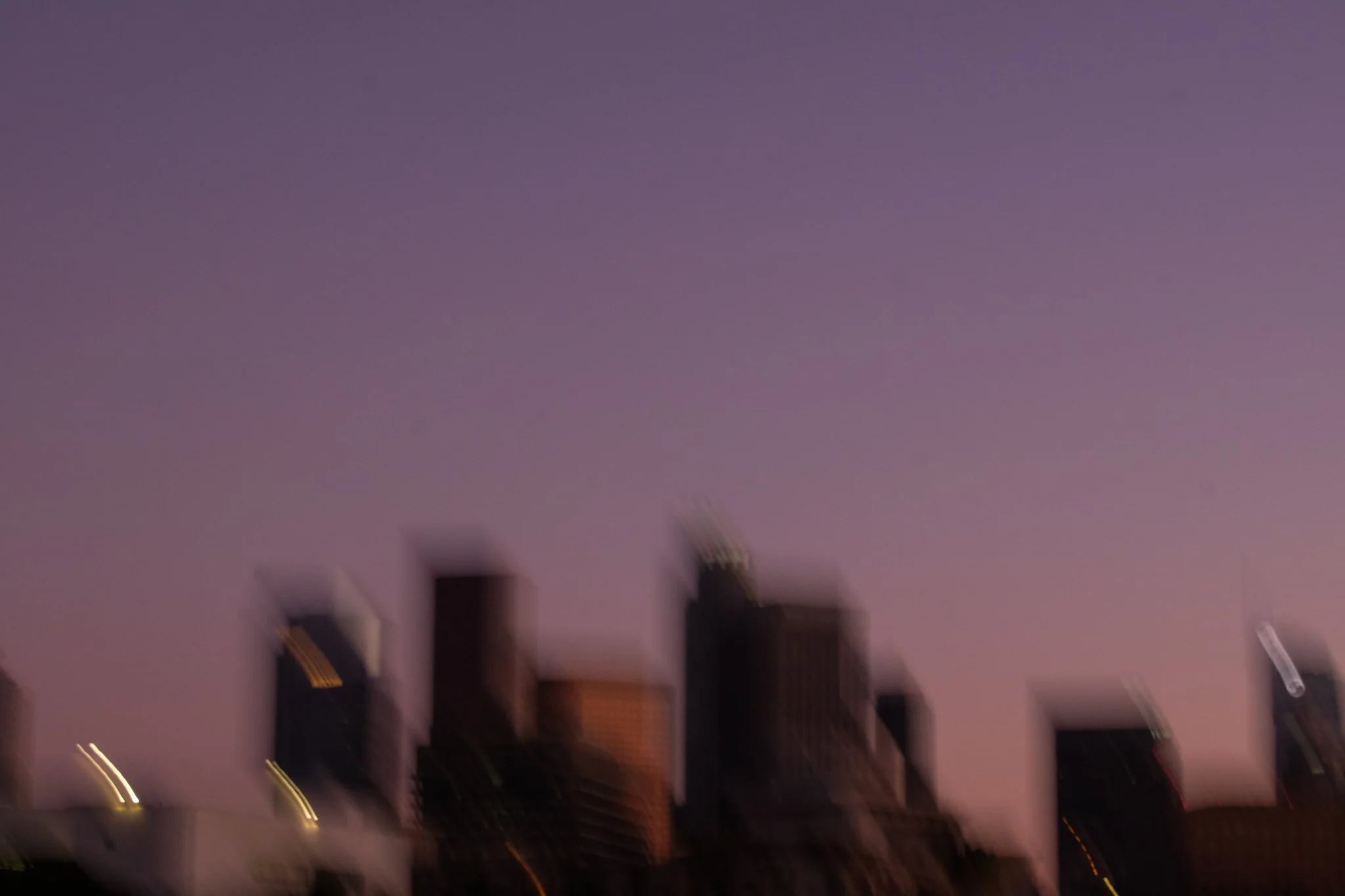 Blurry image of city buildings at dusk or night, with purple sky and some lights visible.