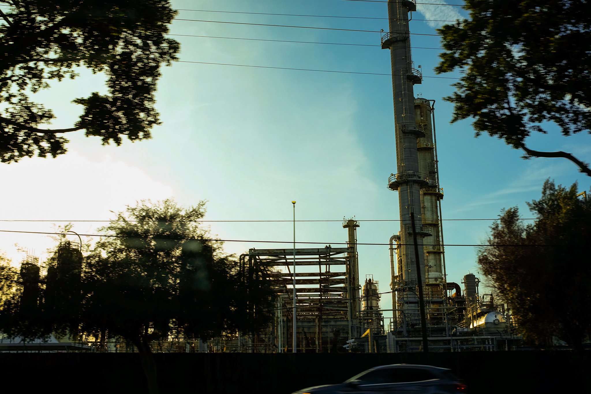 Industrial plant with tall smokestacks and piping, surrounded by trees, under a blue sky during daytime.